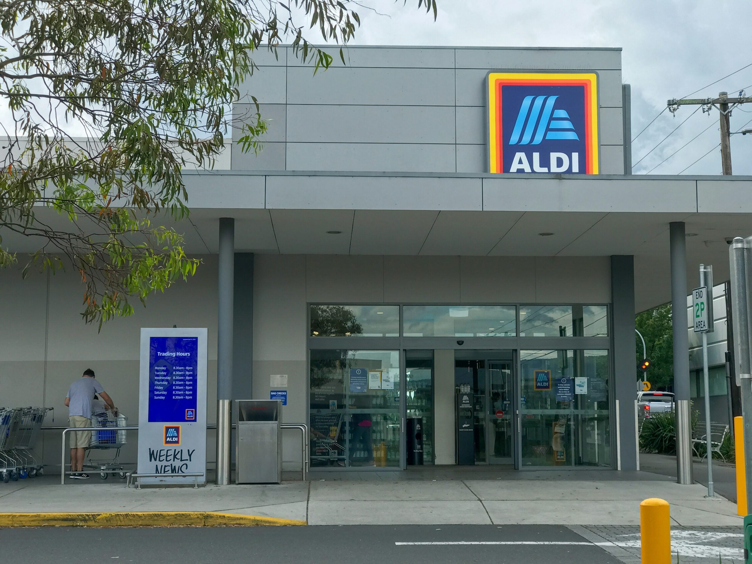 The front entrance of an ALDI supermarket, with glass doors, a large ALDI sign above, shopping carts to the side, and a person unloading groceries near the entrance.