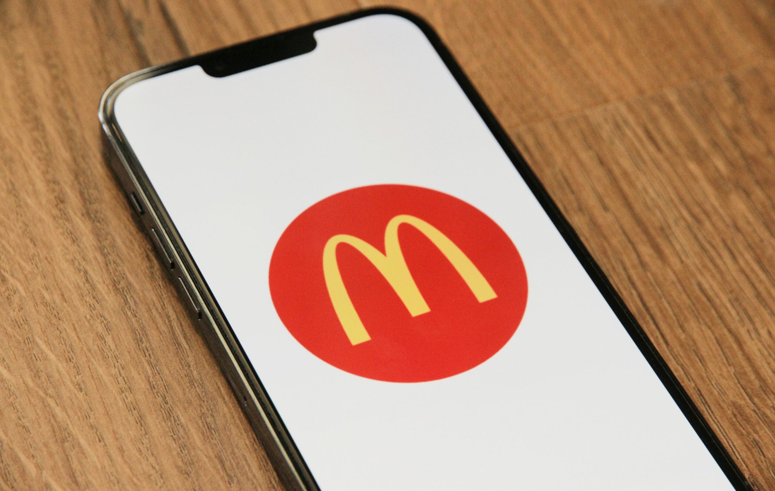 A smartphone displaying the McDonald’s yellow “M” logo inside a red circle on its screen, resting on a wooden surface.