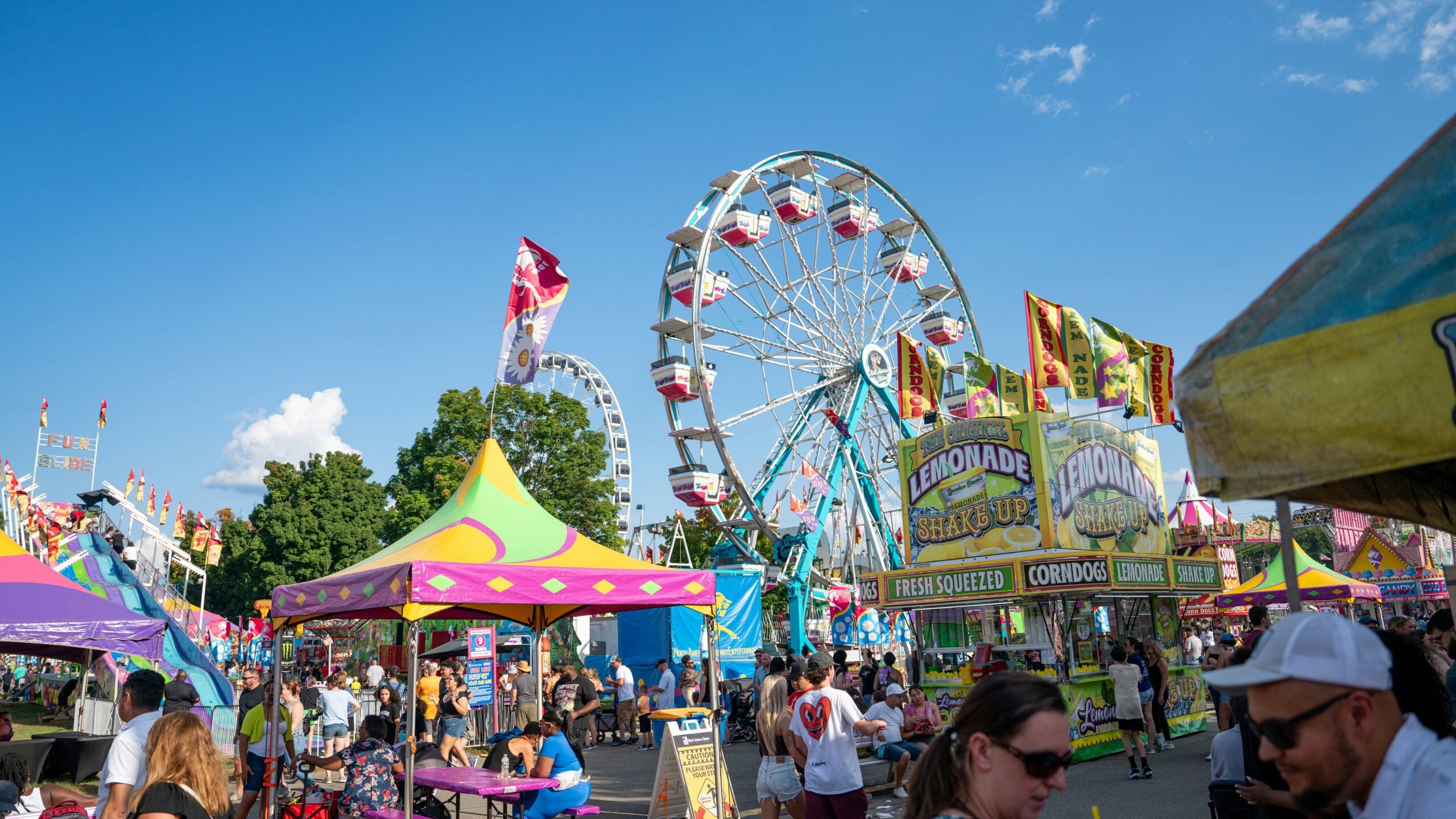 A lively fairground scene with crowds, colorful tents, food stands selling lemonade and corn dogs, tall flags, and a large Ferris wheel under a blue sky with scattered clouds.