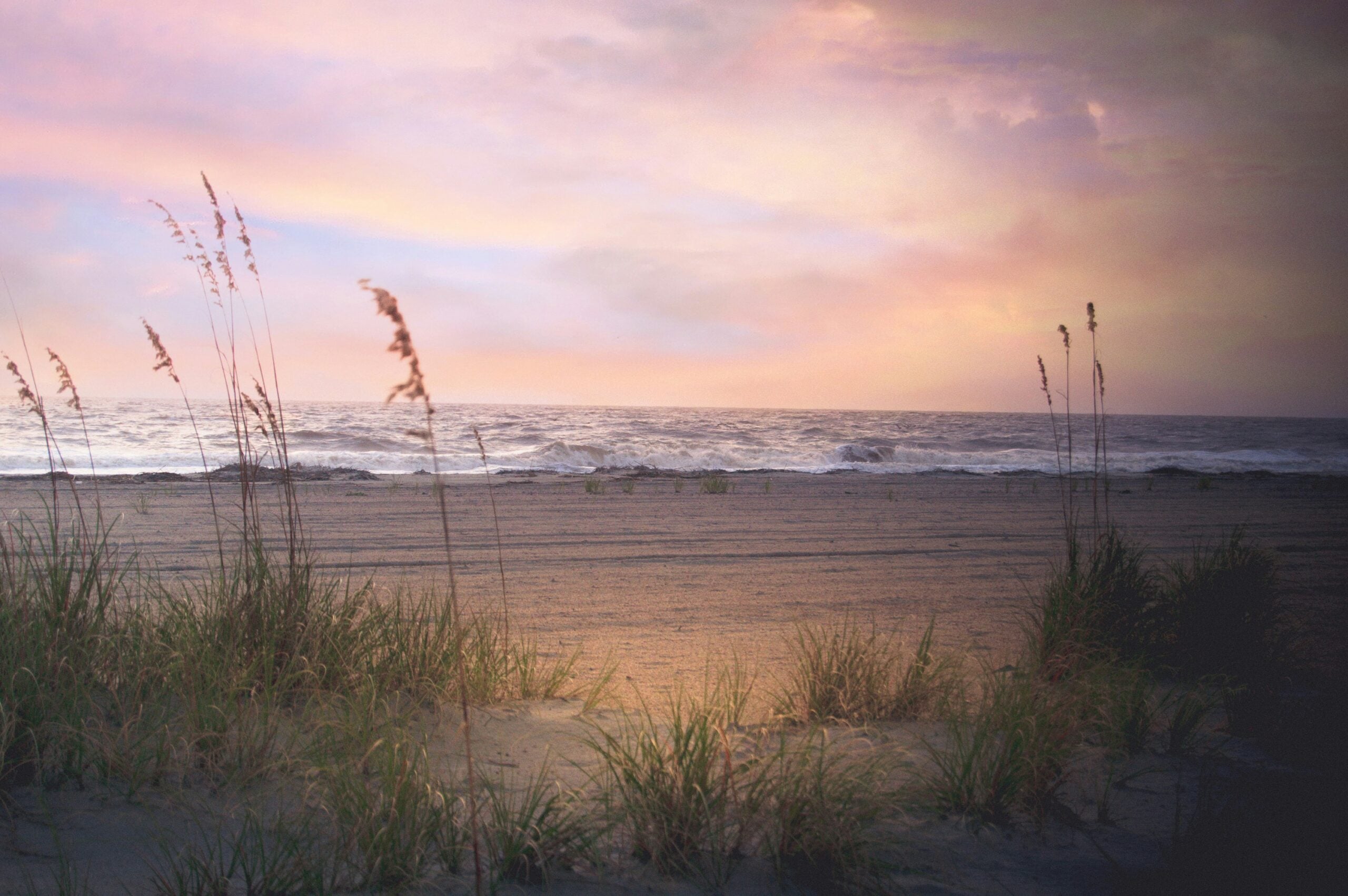 Grassy sand dunes in the foreground frame a tranquil beach with gentle ocean waves under a colorful, pastel sunset sky.