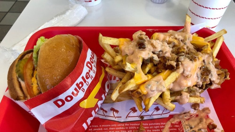 A tray with an In-N-Out Double-Double cheeseburger wrapped in branded paper and a serving of Animal Style fries topped with cheese, grilled onions, and sauce, with drink cups in the background.
