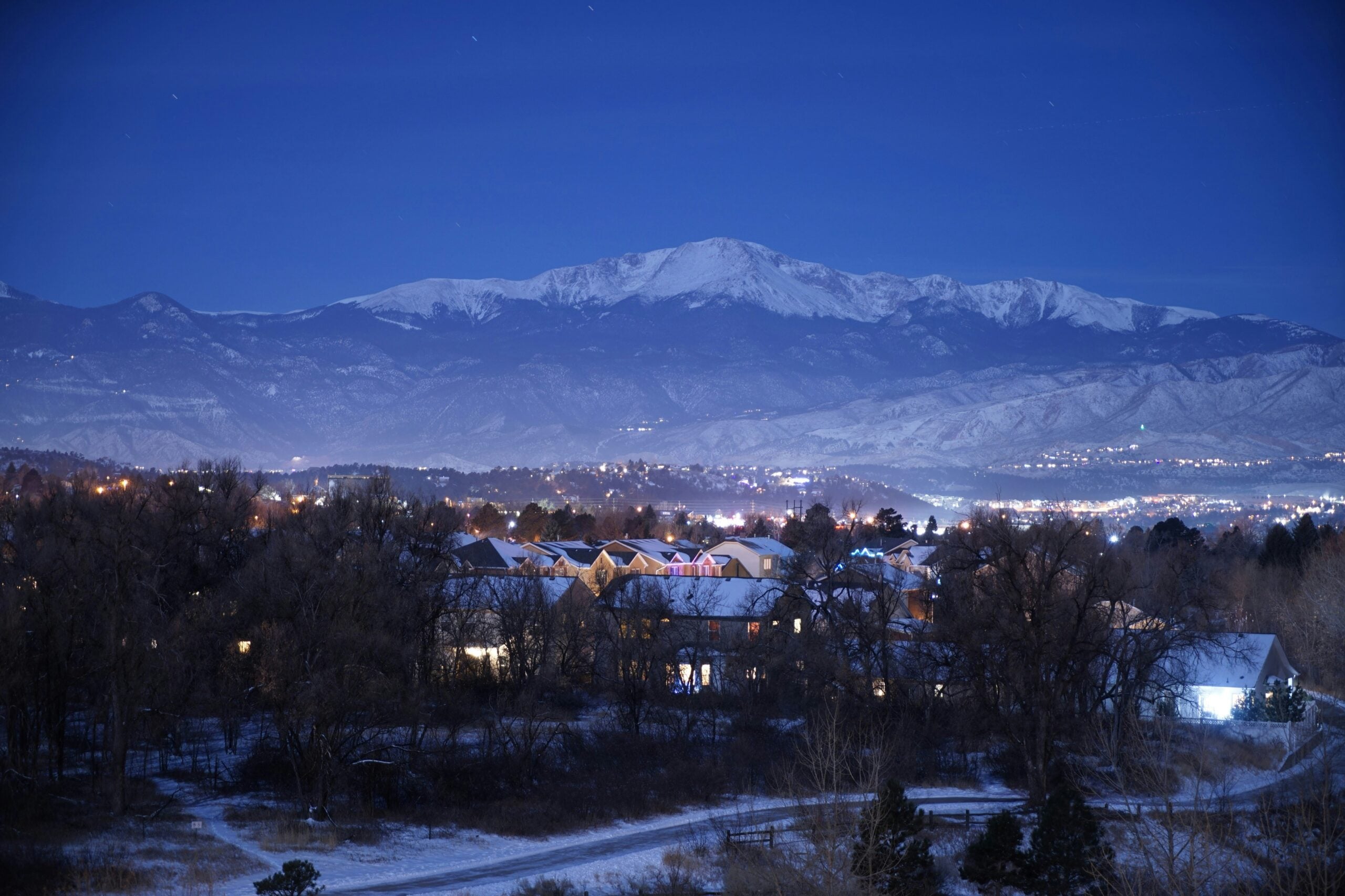 A snowy neighborhood at night is illuminated by warm streetlights, with a large mountain range rising in the background under a clear, dark blue sky.