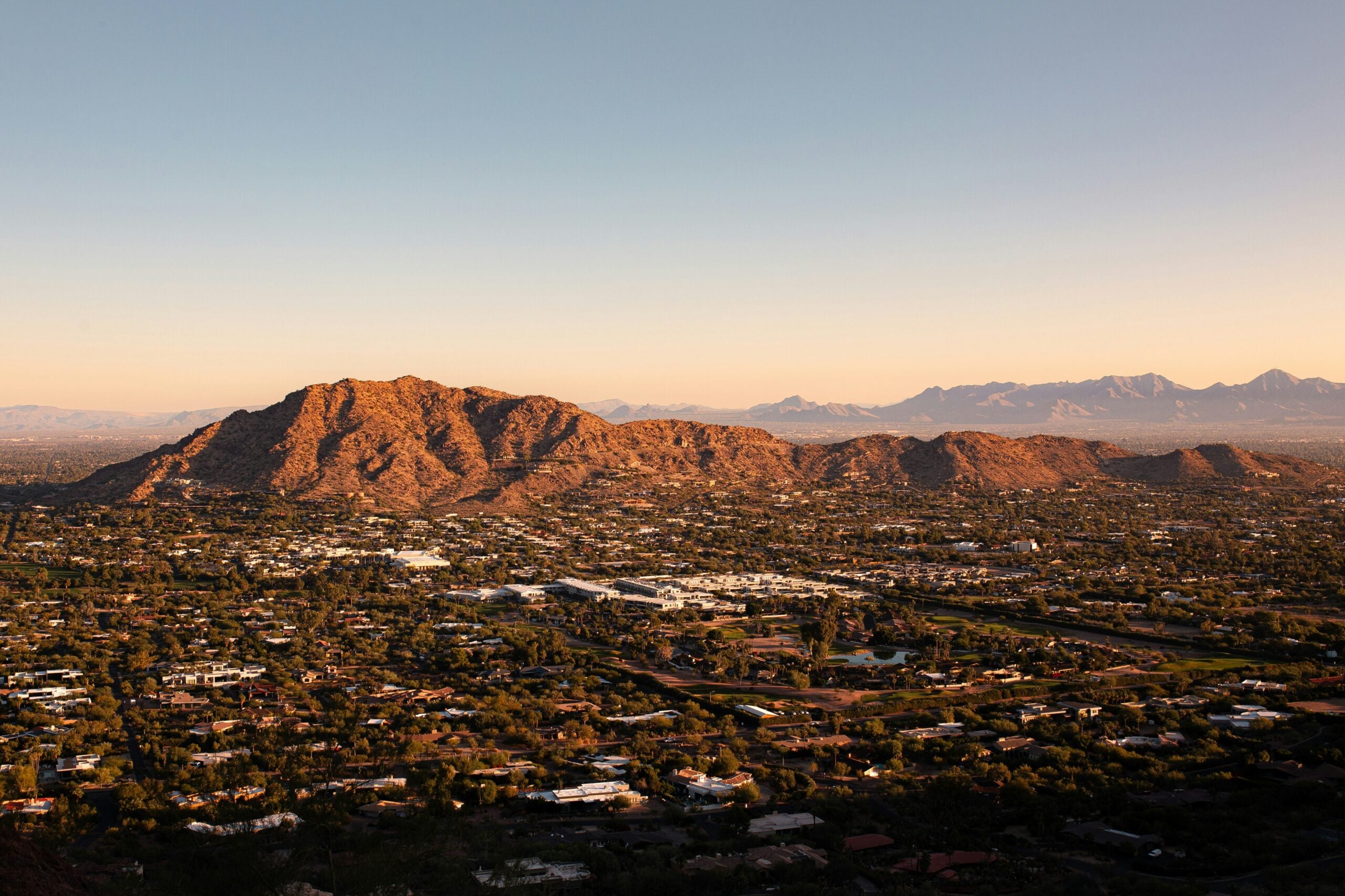 A wide view of a city with scattered buildings, green vegetation, and a large, rugged mountain in the background under a clear sky at sunset.