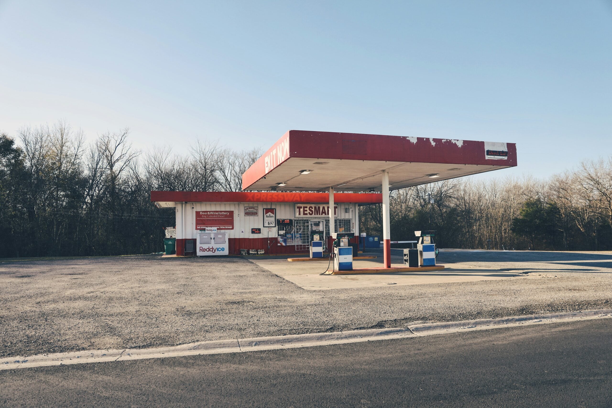 An old, small gas station with a red and white canopy and pumps stands alone in a mostly empty parking lot, surrounded by leafless trees under a clear blue sky.