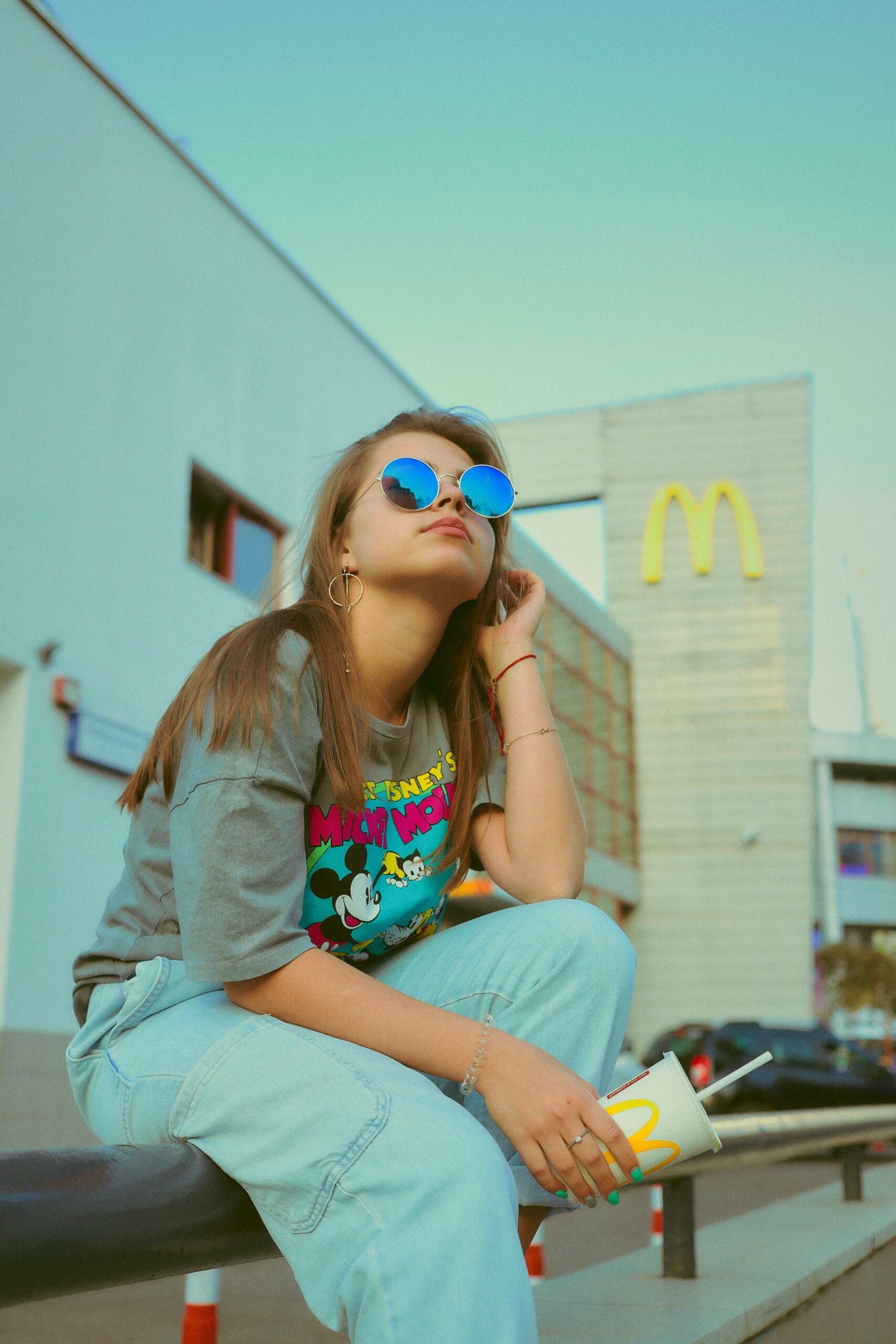 A young woman wearing blue sunglasses and a Mickey Mouse t-shirt sits on a bench outside a McDonald’s, holding a McDonald’s cup, with the restaurant’s golden arches sign visible in the background.