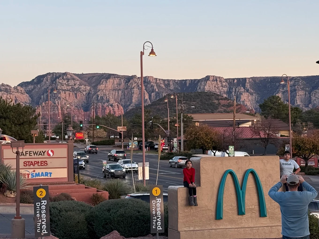 Two children sit on a large McDonald's sign as someone takes their photo. Behind them is a busy street, shopping centers with Safeway and Staples signs, and scenic rocky mountains in the background at sunset.