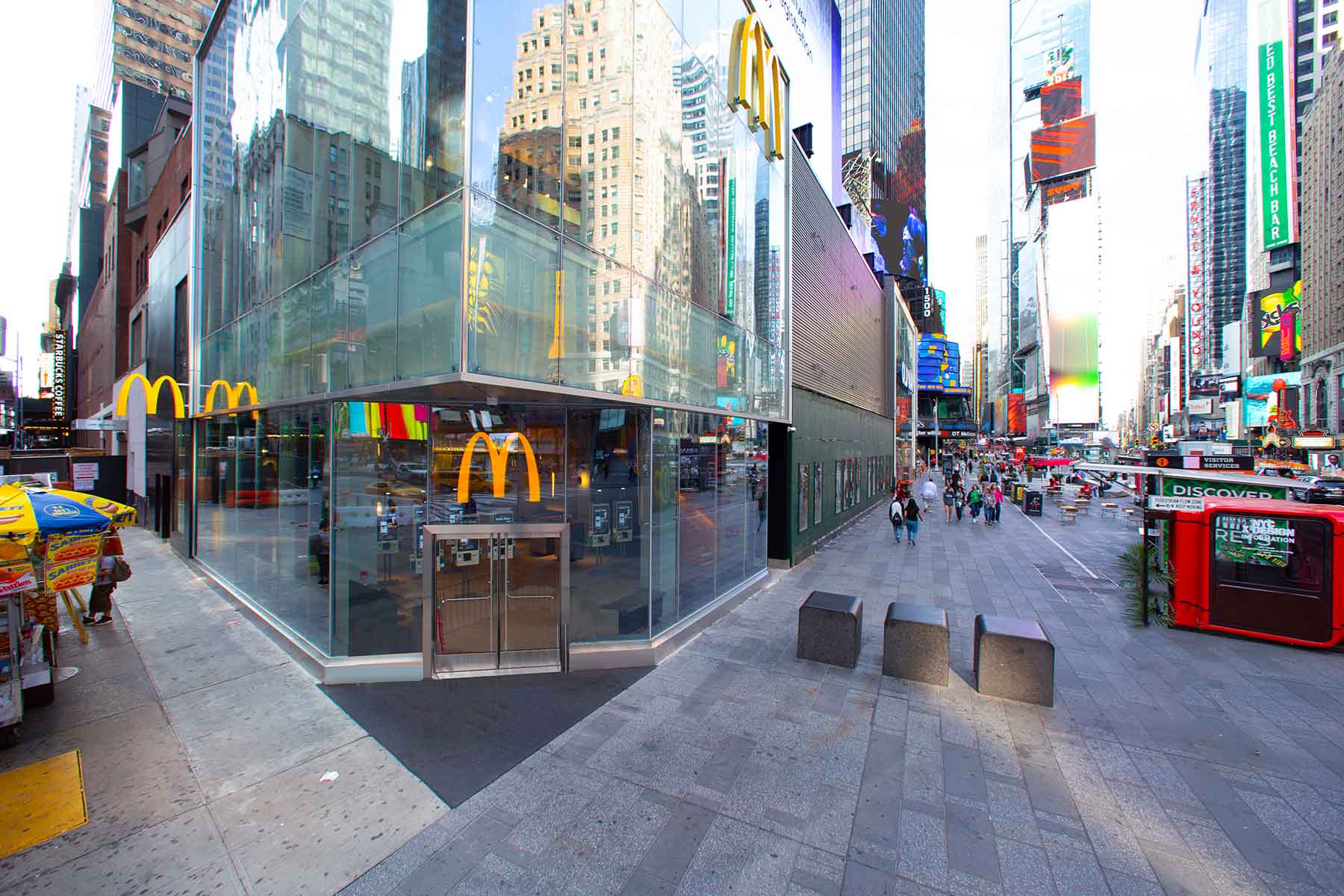 A modern McDonald's restaurant with glass walls on a busy street corner in Times Square, New York City, surrounded by people, street vendors, and bright digital billboards.