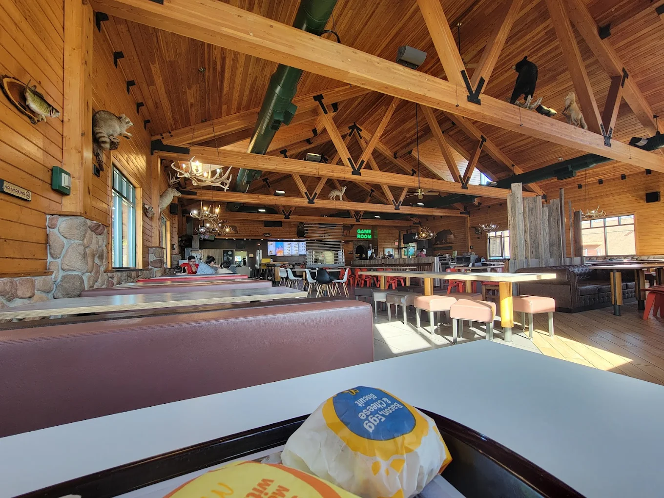 A spacious, rustic-style restaurant with wooden beams, animal decor on the walls, and antler chandeliers. Booths and tables fill the room. In the foreground, wrapped breakfast sandwiches sit on a white table. McDonald's locations.