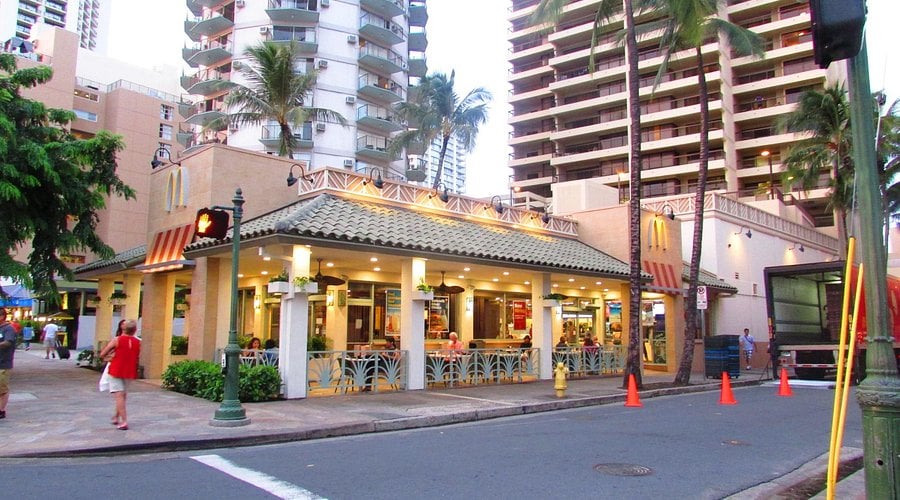 A McDonald’s restaurant with outdoor seating and tropical plants on a city street, surrounded by tall palm trees and high-rise buildings, with people walking nearby. Orange cones line the street in front.