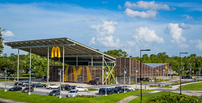 A modern McDonald’s restaurant with a large solar panel roof, numerous parked cars, and green trees in the background under a blue sky with clouds.