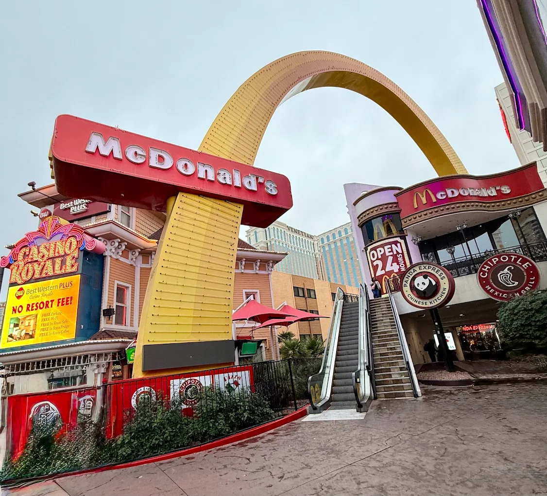 A McDonald’s with a large yellow arch, escalator, and signs, next to Casino Royale and Chipotle, under a cloudy sky on a busy city street.