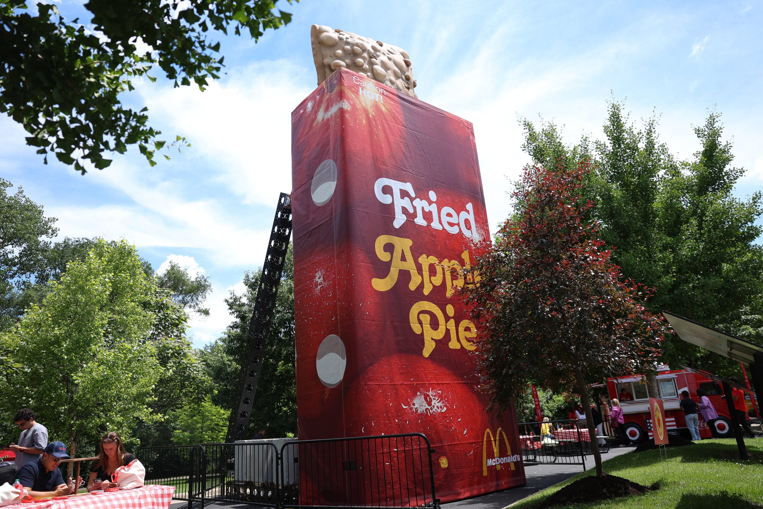 A giant display shaped like a McDonald's fried apple pie stands outdoors, featuring bold "Fried Apple Pie" text. Nearby are trees, a picnic area with people, and a red food truck. The sky is clear and sunny.