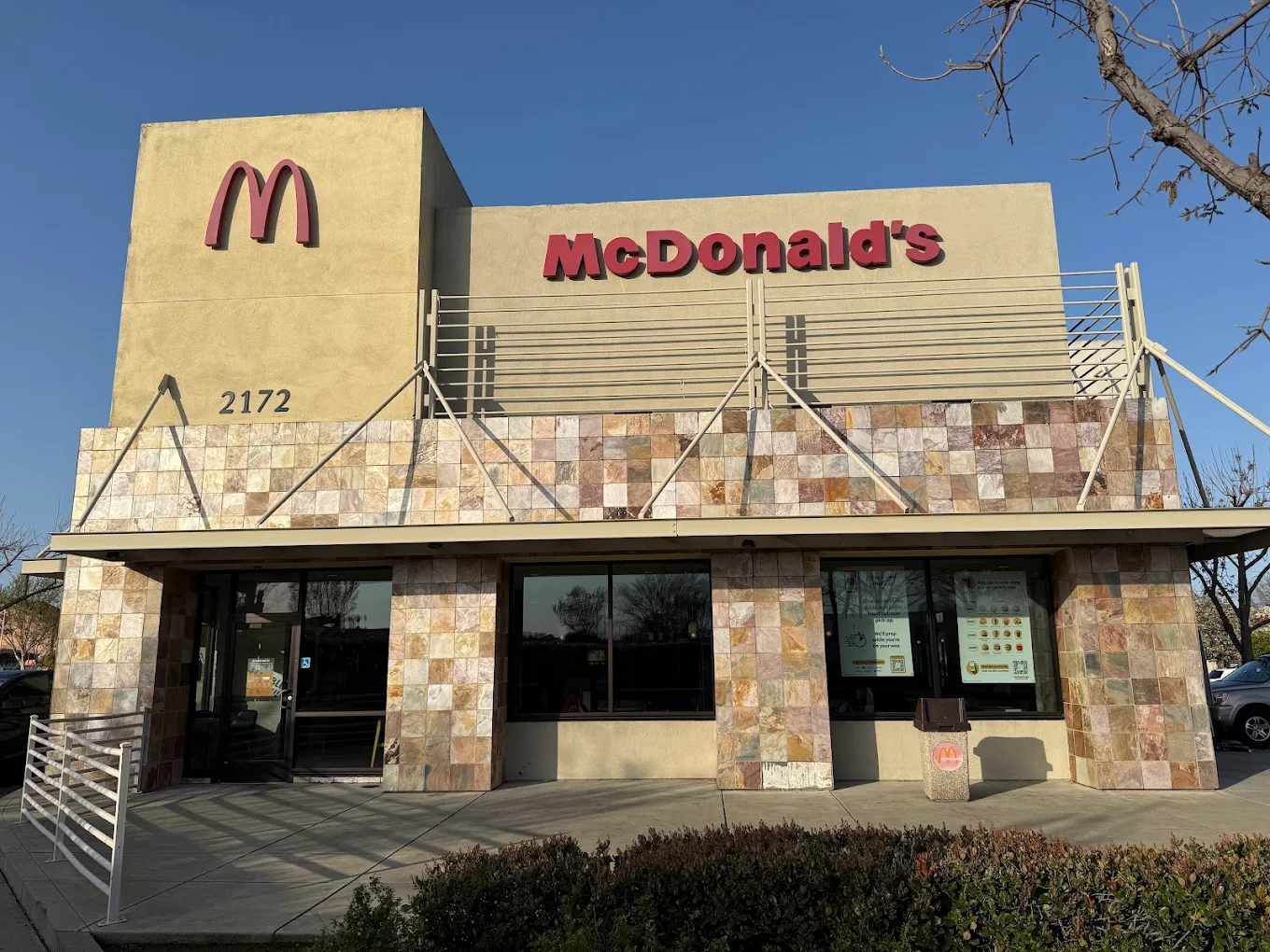 A McDonald's restaurant with a tan and stone-tiled exterior, large McDonald's logo, and the address number 2172 on the building. The sky is clear and trees are visible in the reflection of the windows.