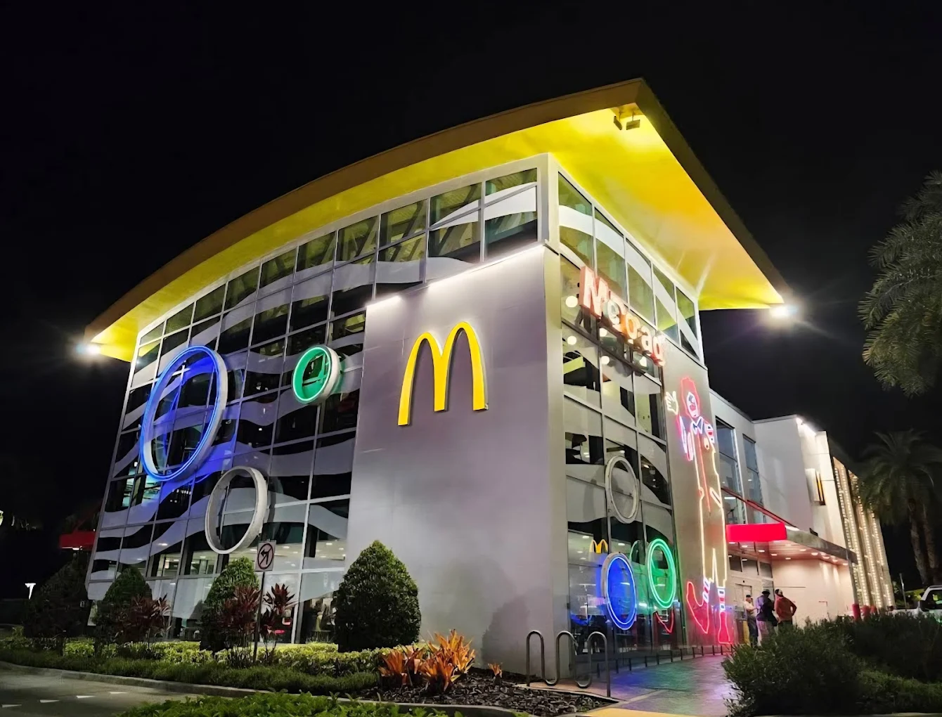 A modern McDonald's restaurant at night, featuring large glass windows, colorful neon lights, and the yellow McDonald's "M" logo on a white wall with a distinctive yellow roof. Some greenery is visible in front.