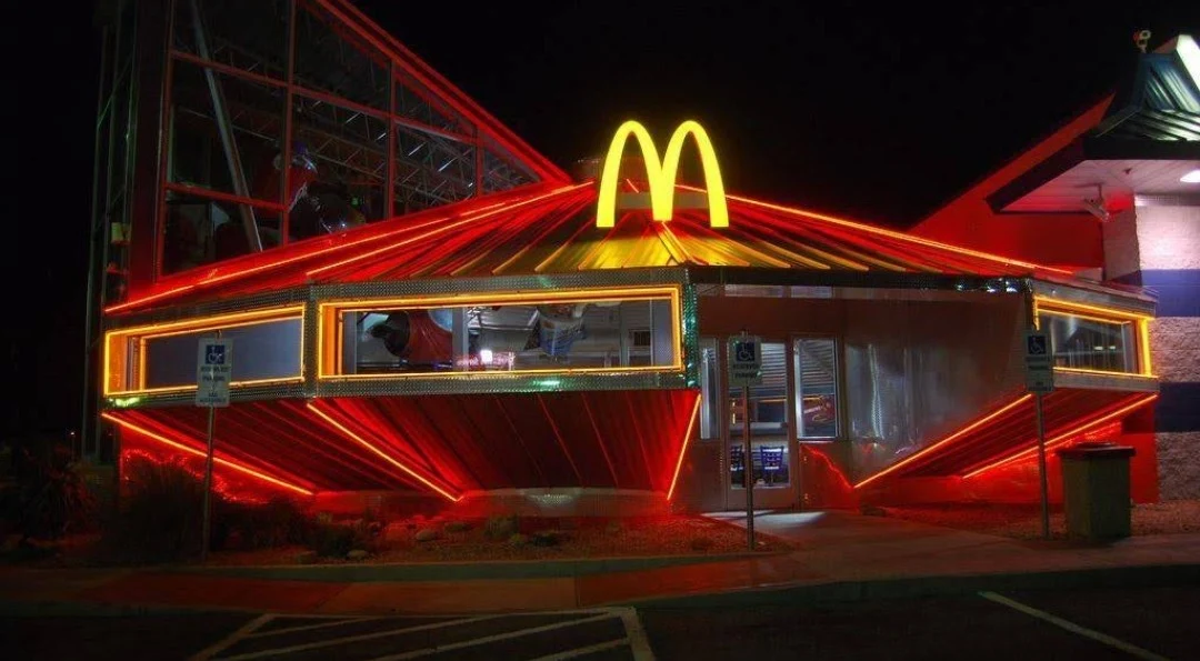 A McDonald's restaurant at night with a bright yellow "M" logo and vibrant red and orange neon lights outlining the building, giving it a futuristic, spaceship-like appearance.