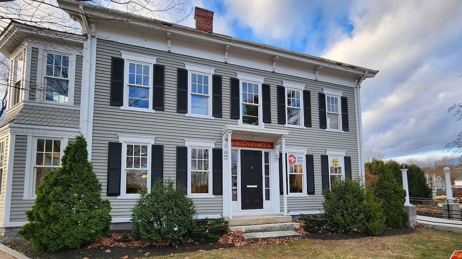 A gray two-story historic house with black shutters has a McDonald's sign above the door, blending classic architecture with modern fast food branding. Shrubs and a pathway are in front, under a partly cloudy sky.