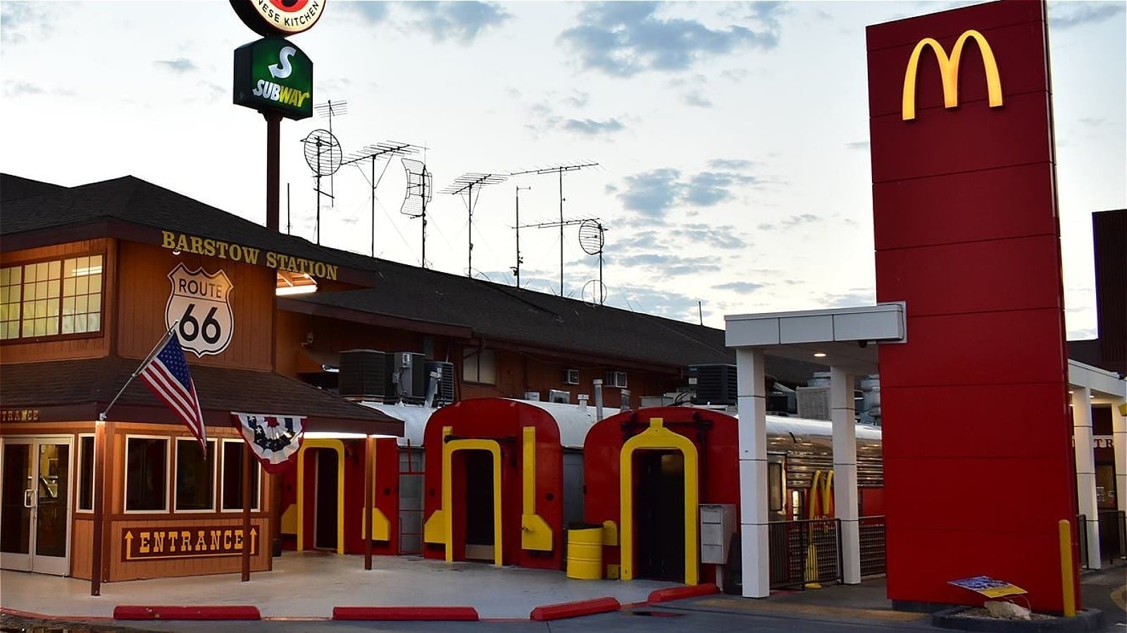 Barstow Station on Route 66 featuring a Subway sign, a McDonald's sign, a brown building with a Route 66 emblem, and colorful, train car-shaped entrances under a partly cloudy sky.