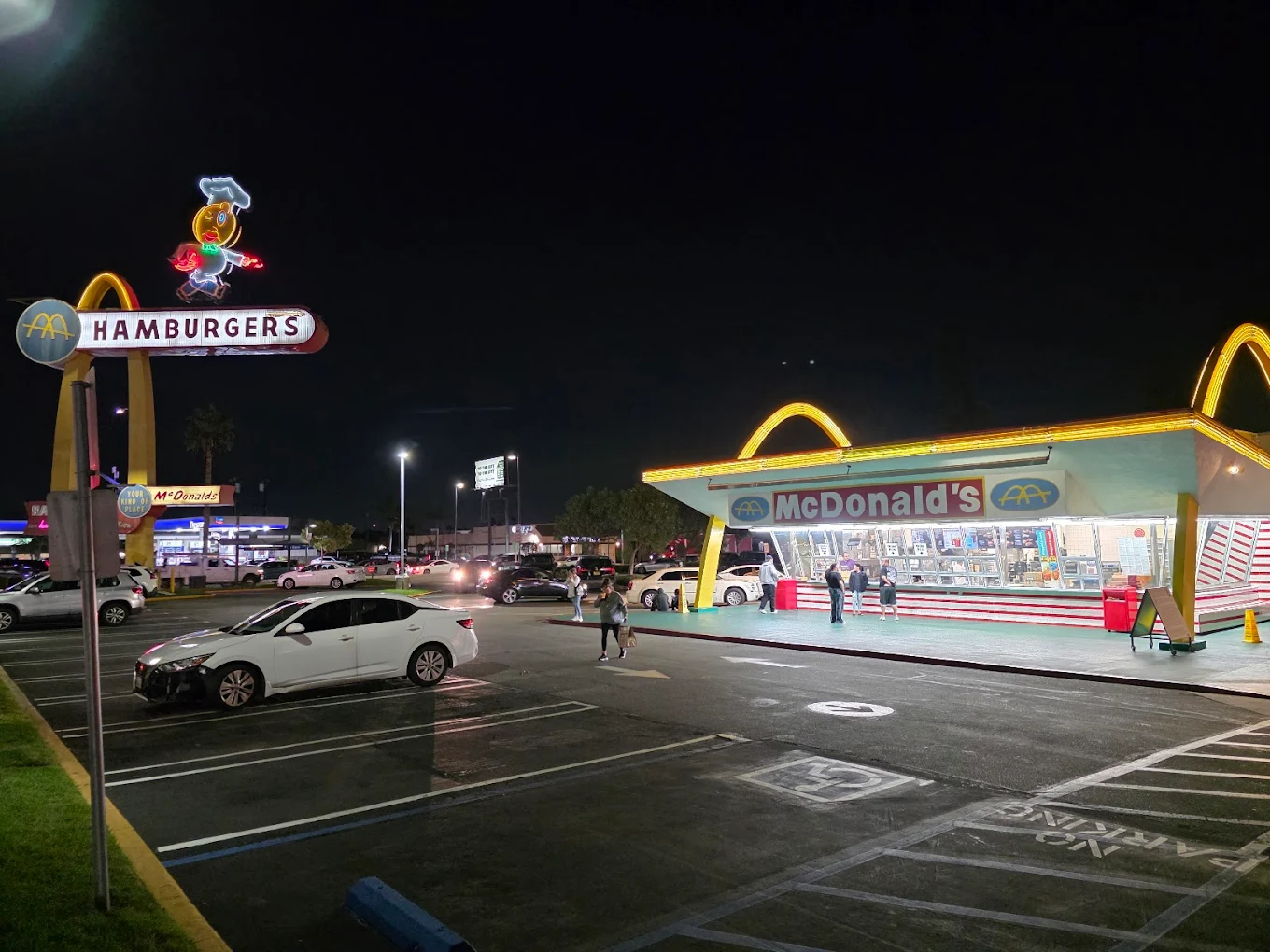 A brightly lit, retro-style McDonald's at night features golden arches, a vintage "HAMBURGERS" sign, and people walking in the parking lot with cars parked nearby. Other businesses are visible in the background.