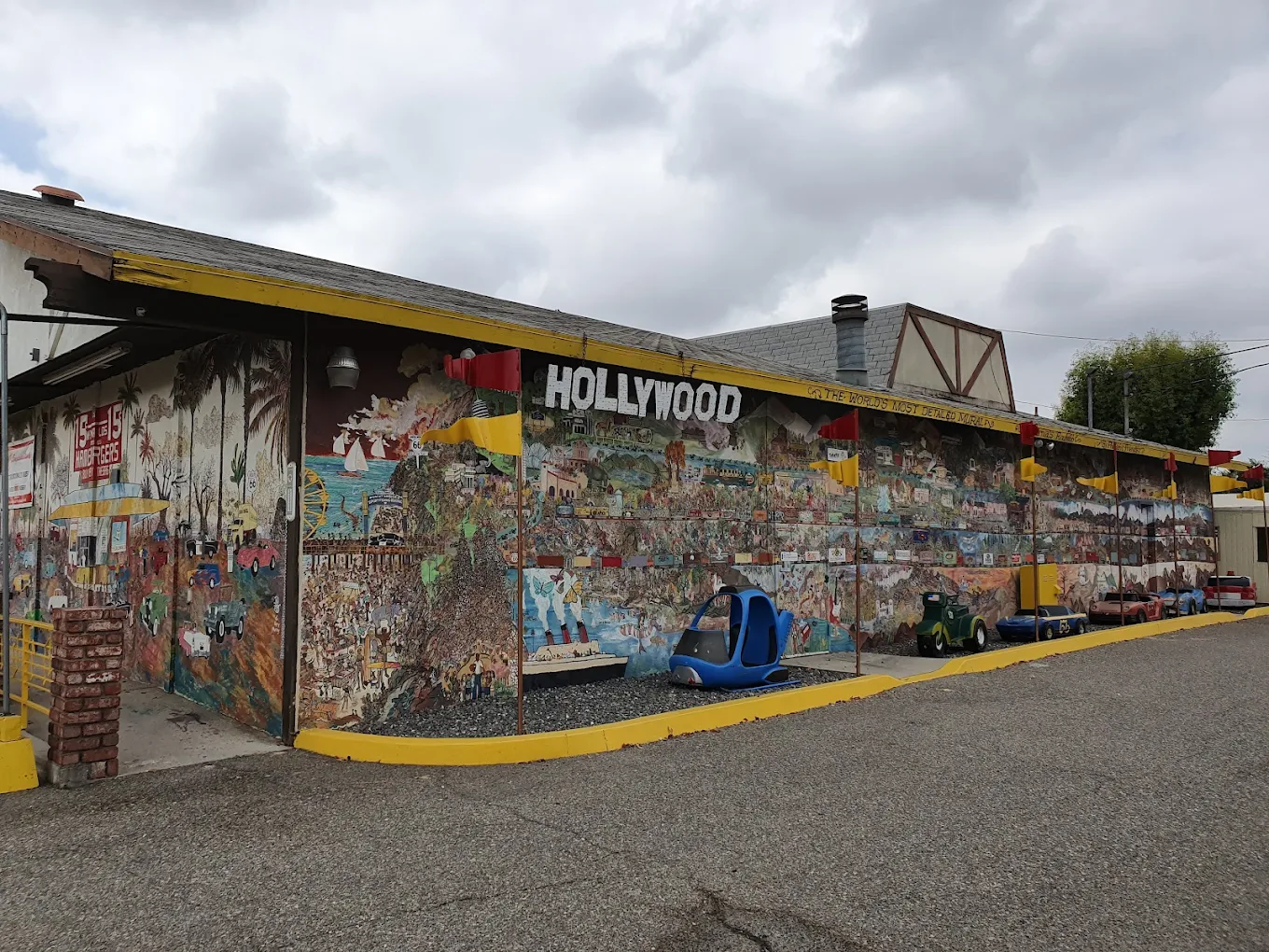 A building with a large colorful mural depicting various scenes, topped by a "HOLLYWOOD" sign. Small blue and green go-karts are parked beside it. The sky is cloudy, and the ground is paved.