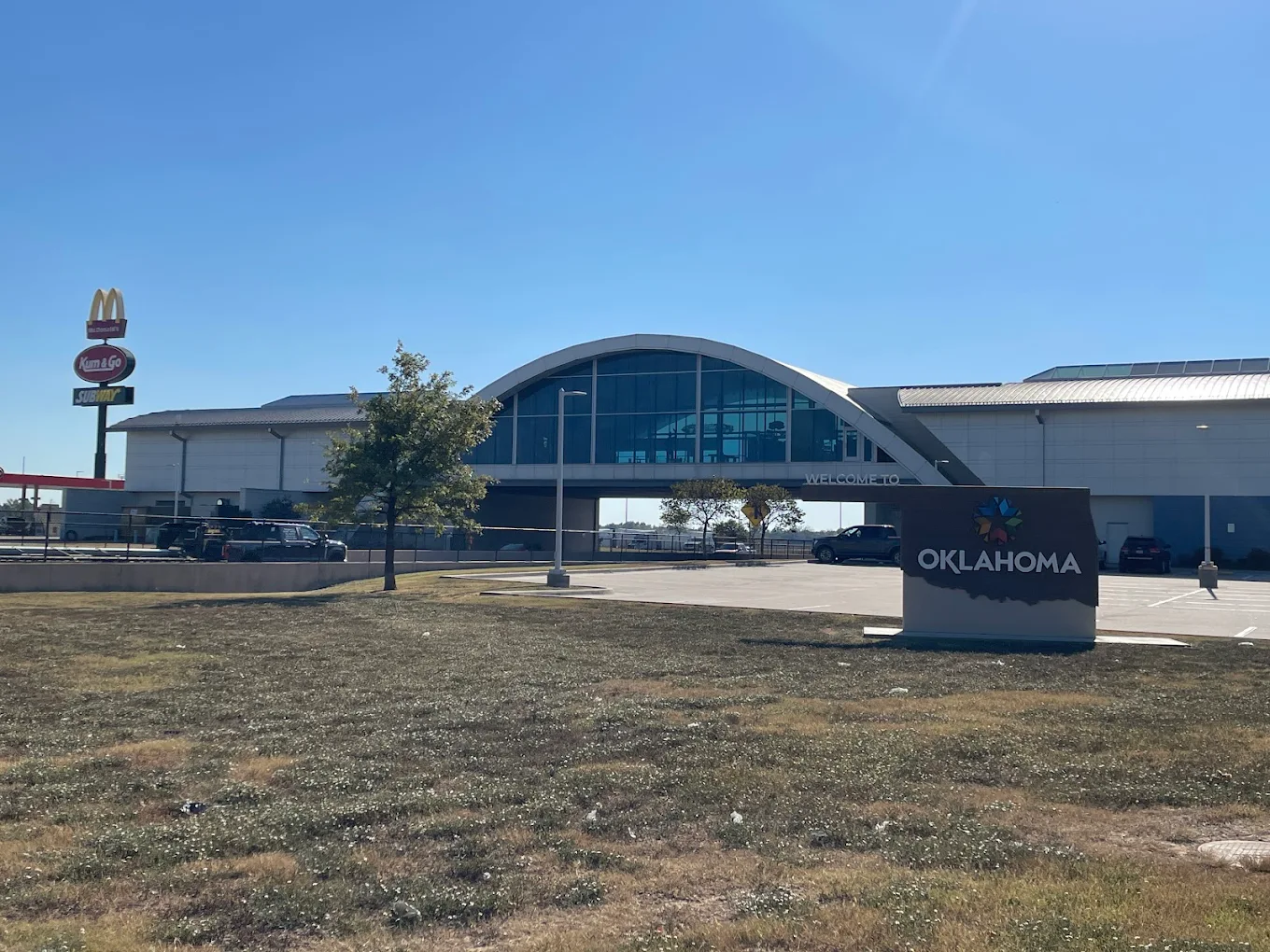 A modern building with a curved roof and large windows stands behind a sign that reads "Oklahoma." A McDonald's sign is visible to the left, and cars are parked in the lot under a clear blue sky.