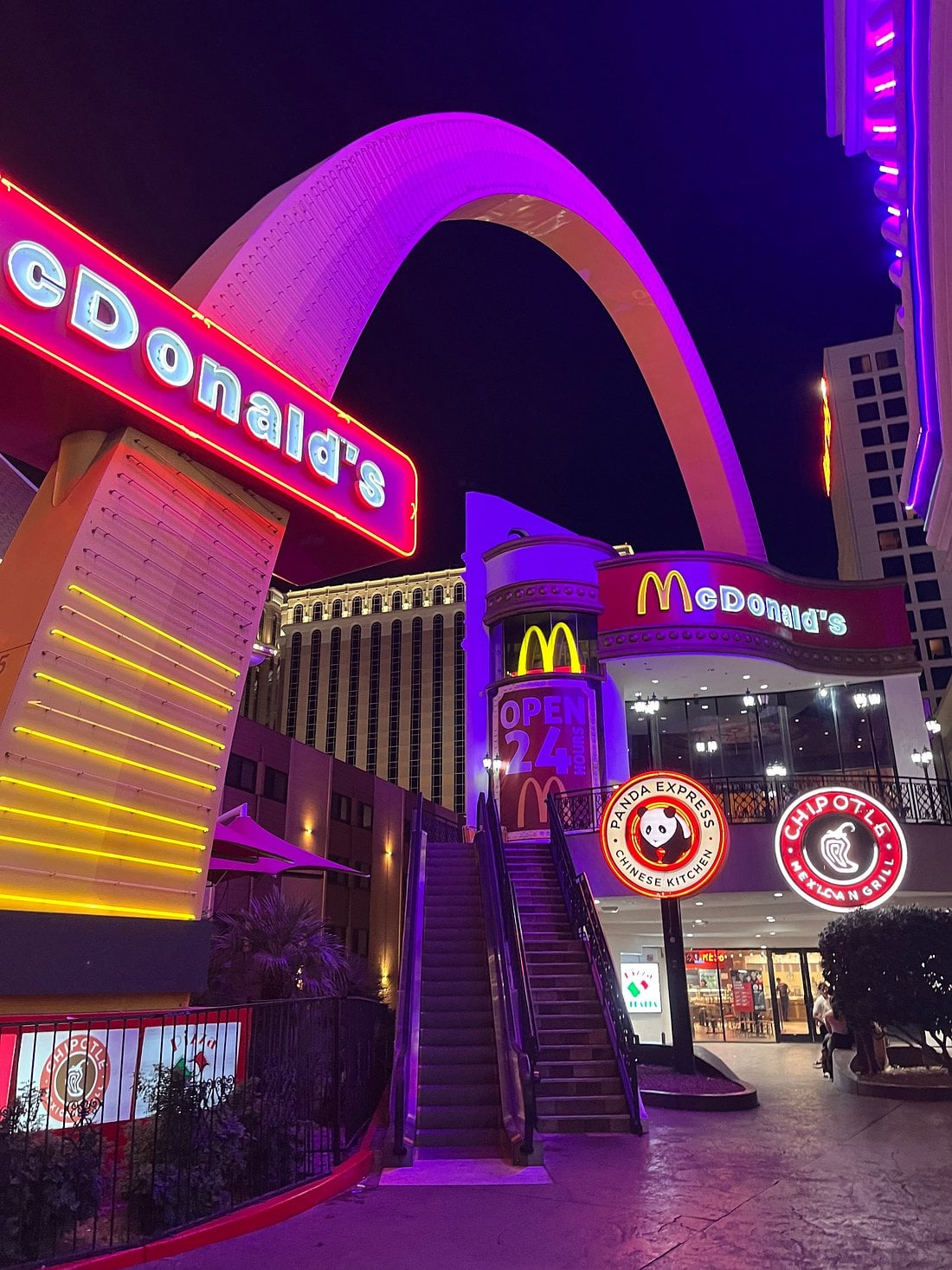A brightly lit McDonald’s with neon lights at night, featuring a large golden arch above the building. Among the coolest McDonald's locations, it sits near Panda Express and other restaurants, with stairs leading to its entrance.
