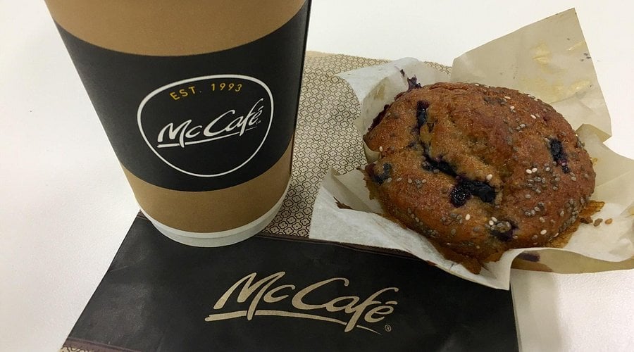 A McCafé coffee cup sits next to a blueberry muffin on a napkin with the McCafé logo. The muffin is partially unwrapped, revealing blueberries and sprinkled sugar on top.