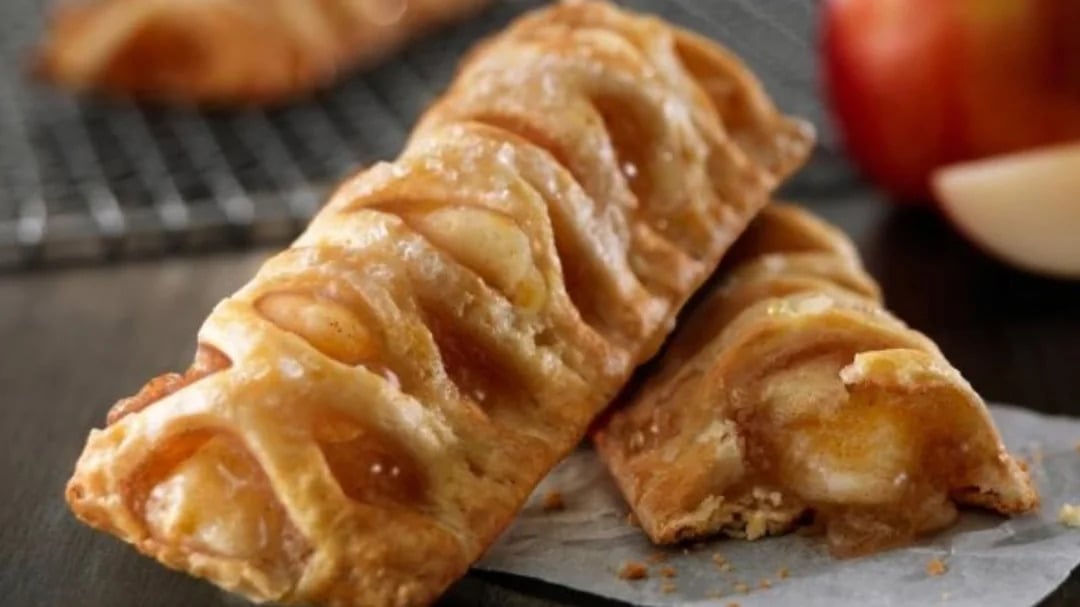 Two golden-brown apple turnovers with flaky pastry and visible apple filling, one resting on top of the other—a tempting take on classic fast-food desserts—with a blurred apple and cooling rack in the background.
