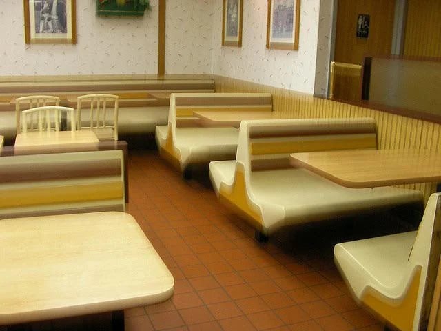 Empty fast food restaurant interior with beige and yellow booths, wooden chairs, and tiled floor. The walls have light wallpaper and framed pictures, creating a clean, quiet atmosphere.