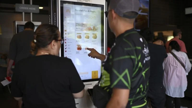 Two people stand in front of a McDonald's self-service kiosk, looking at the menu and making a selection. Other customers are visible in the background, waiting in line.