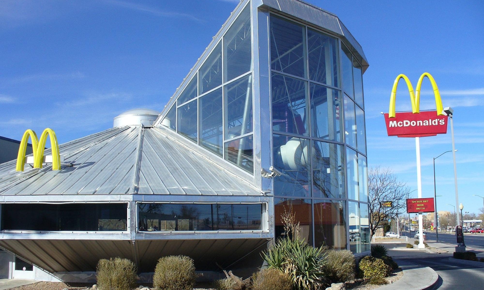 A McDonald’s restaurant with a unique, modern design featuring large glass walls and metal siding. This location is one of the Coolest McDonald's Locations, with two yellow arches and a red sign outside under a clear blue sky, framed by shrubs.