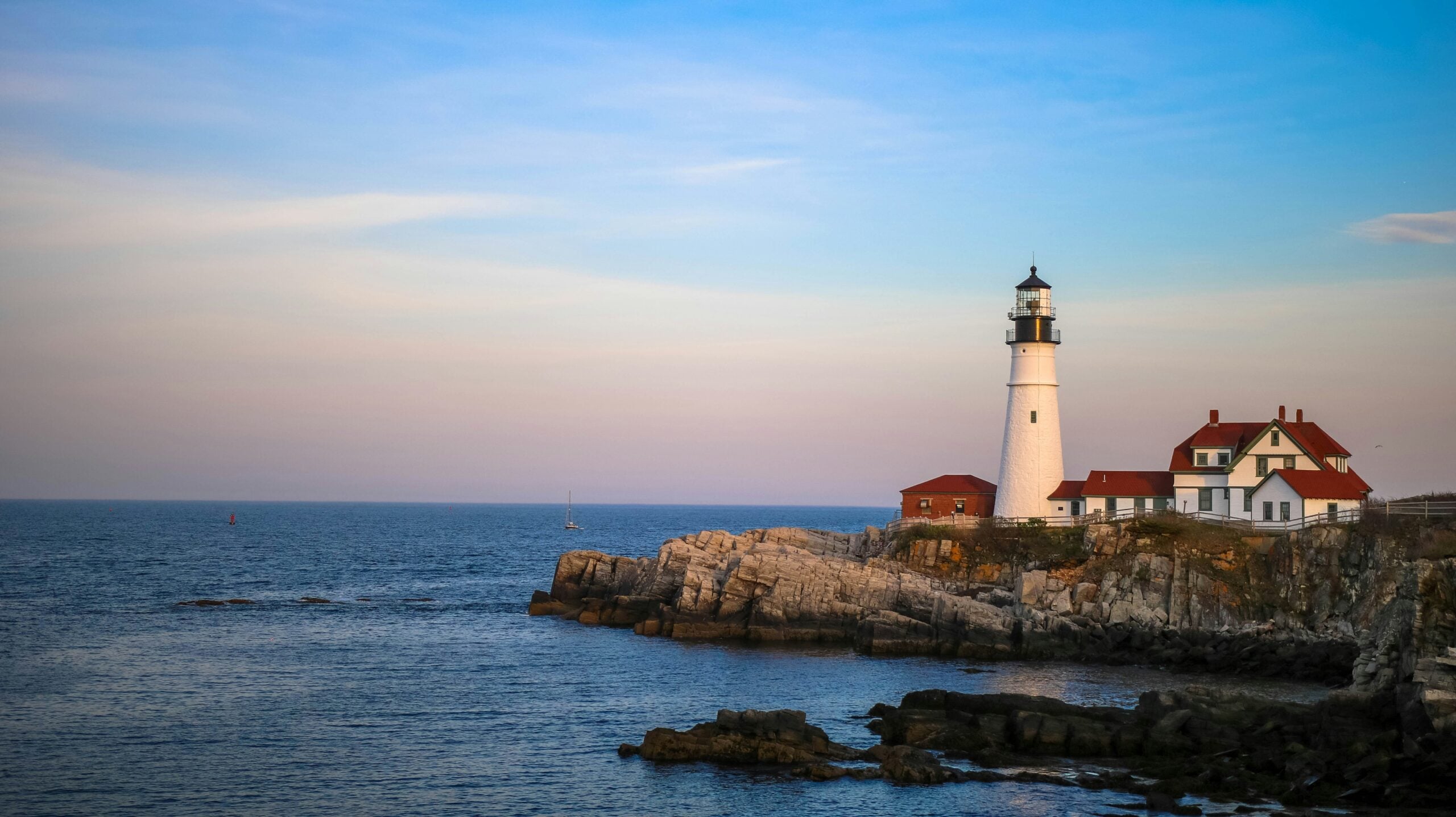 A white lighthouse with a black top stands on a rocky coastline next to white buildings with red roofs, overlooking a calm blue sea under a partly cloudy sky at sunset.