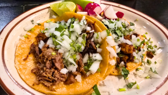 Two tacos topped with chopped onions and cilantro on a plate, with lime wedges and sliced radishes on the side. Enjoy these seasoned meat tacos on soft corn tortillas from our Family-Owned Mexican Restaurant.