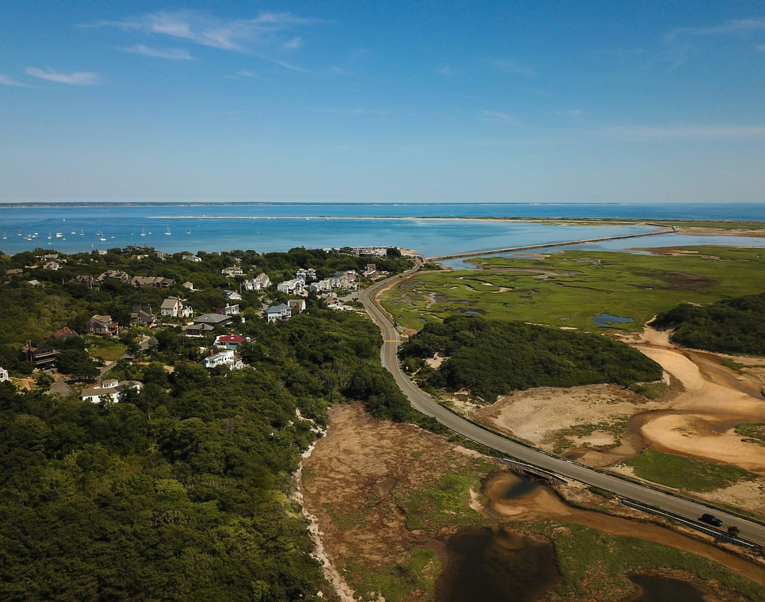 Aerial view of a coastal town with houses, a winding road, green marshlands, and a blue bay dotted with boats under a clear sky.