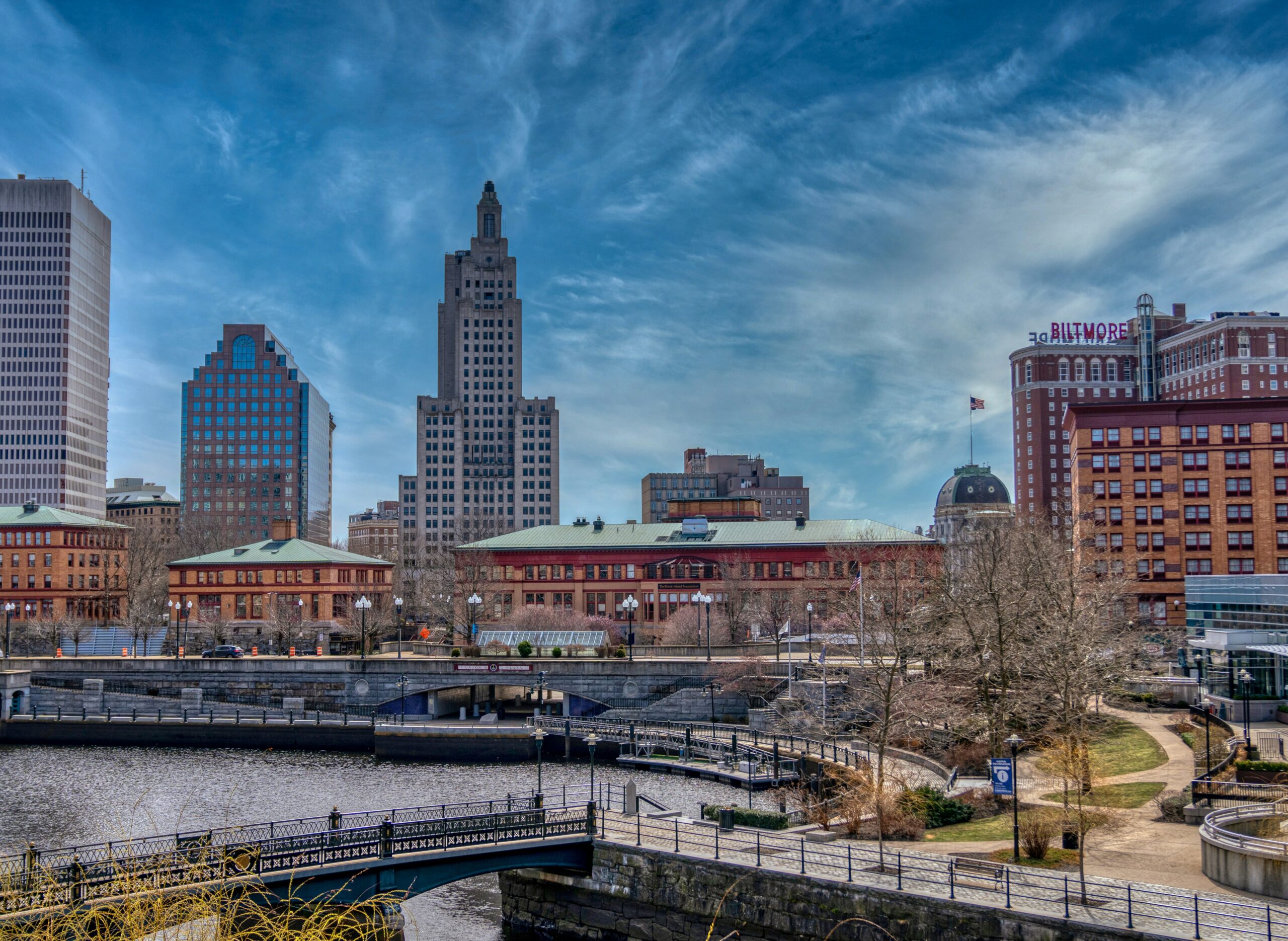 A cityscape featuring a river, pedestrian bridges, and tall buildings under a blue sky with wispy clouds. The Biltmore Hotel sign is visible on a prominent building to the right.