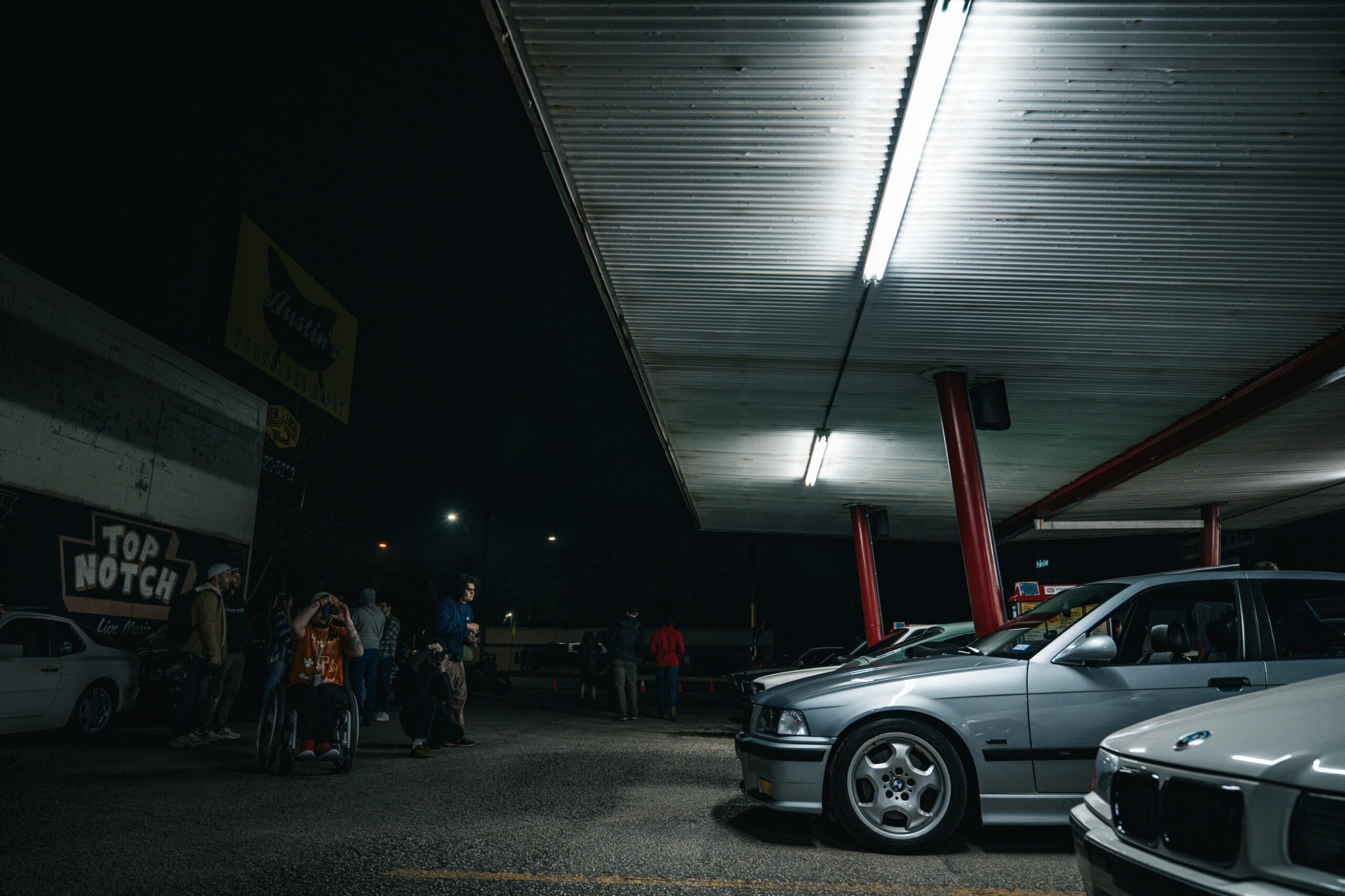 A group of people stand under a brightly lit carport at night, near parked cars, including a silver BMW in the foreground. The scene has a relaxed, urban vibe with a "Top Notch" sign on the nearby building.