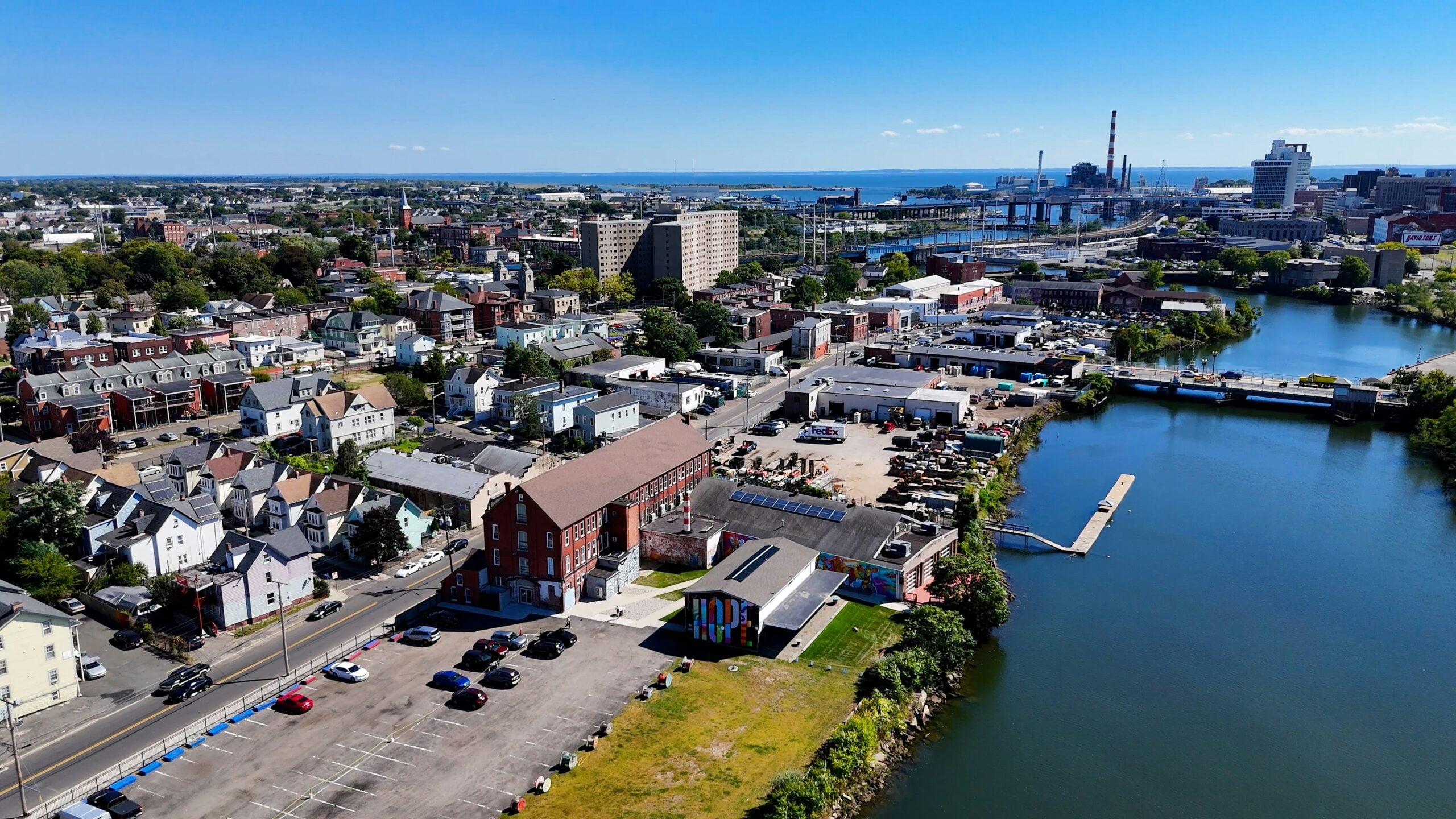 Aerial view of a city beside a river with residential buildings, parking lots, and bridges. Industrial structures are visible in the background under a clear blue sky, highlighting areas benefiting from Cost of Living Relief initiatives.