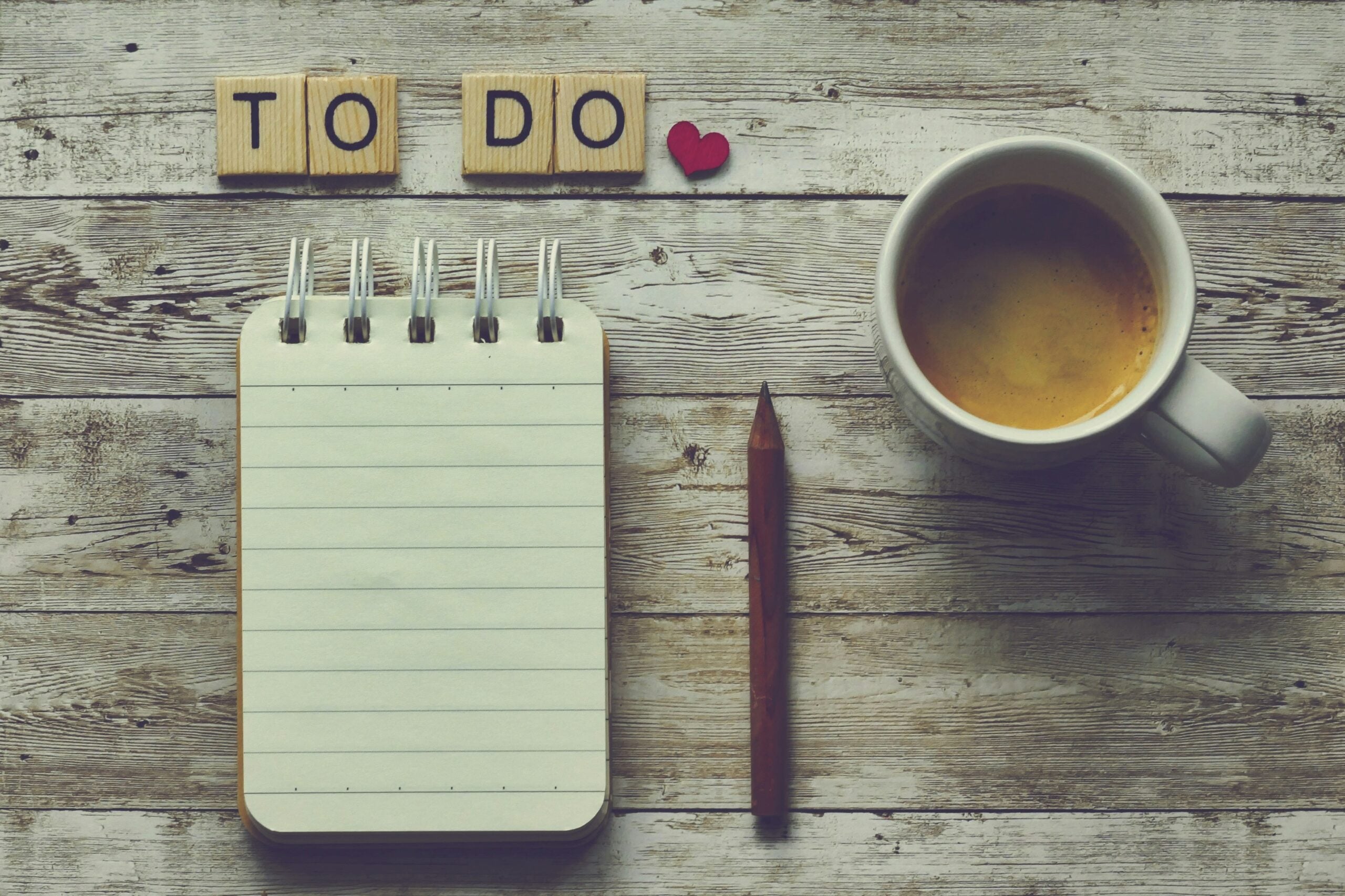 A cup of coffee, a blank notepad with a pencil, and wooden letter tiles spelling “TO DO” with a red heart, all arranged on a rustic wooden surface.