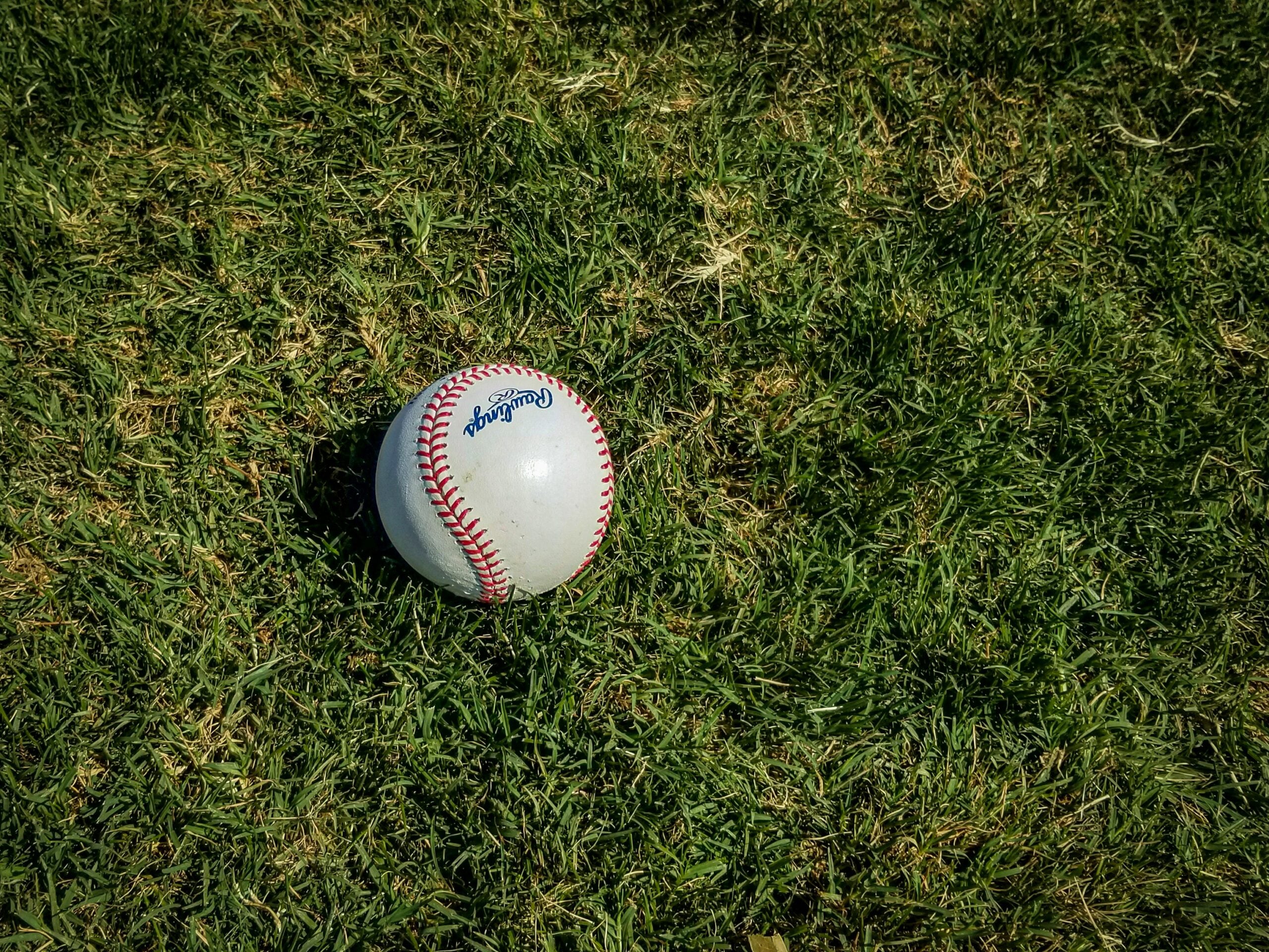 A white baseball with red stitching rests on green grass under sunlight.