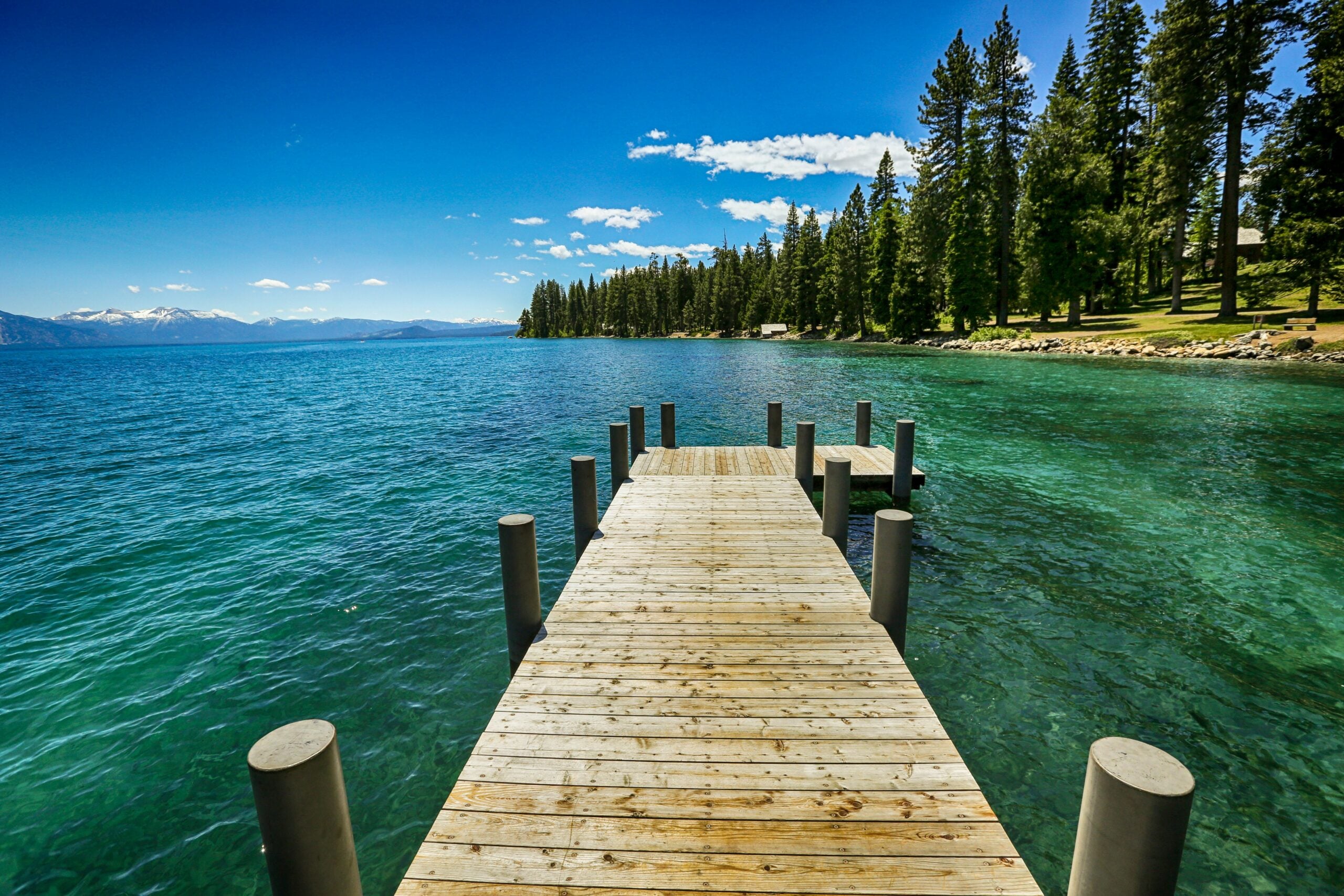 A wooden dock extends over clear blue lake water, surrounded by tall pine trees and a grassy shore under a bright, sunny sky with scattered clouds. Snow-capped mountains are visible in the distance.