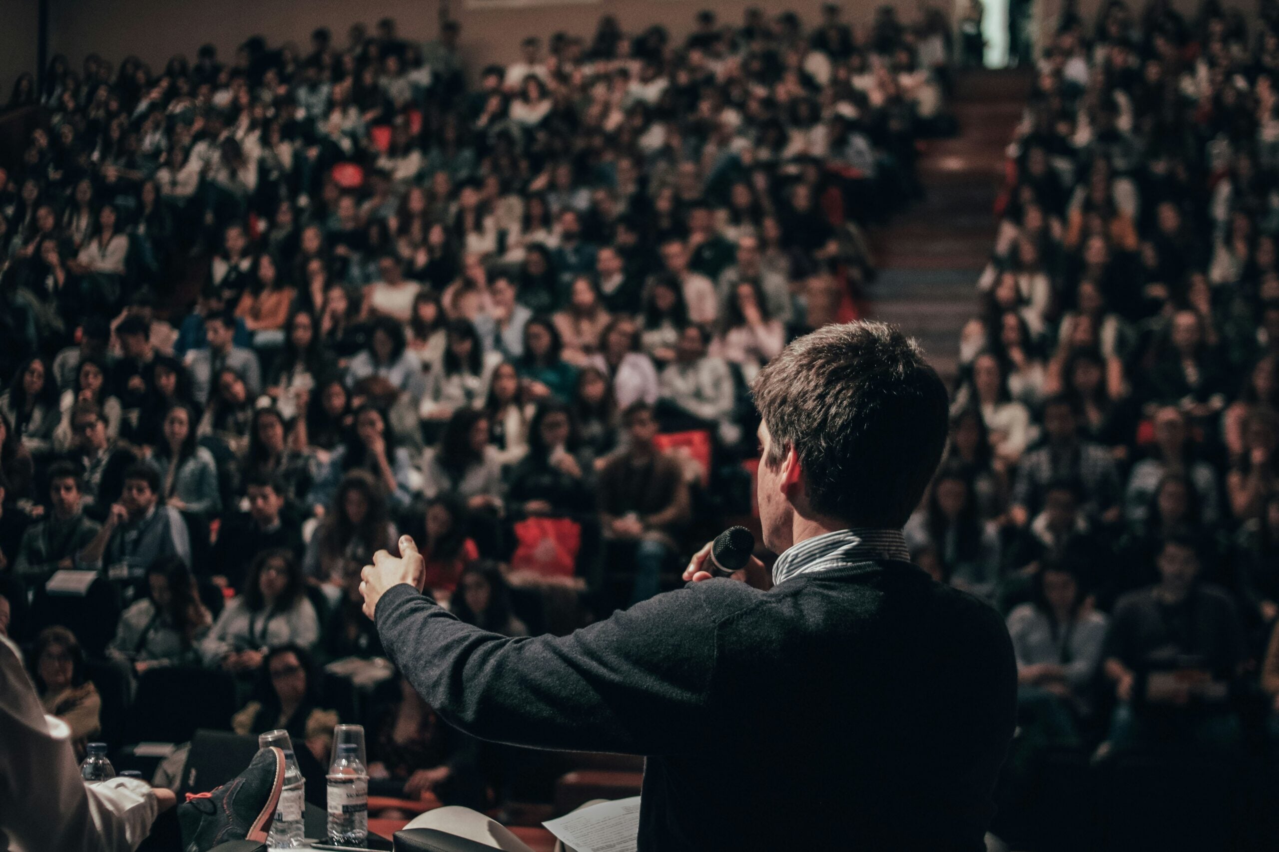 A person holding a microphone speaks to a large, seated audience in an auditorium about Cost of Living Relief. The crowd appears attentive, with the focus on the speaker seen from behind.