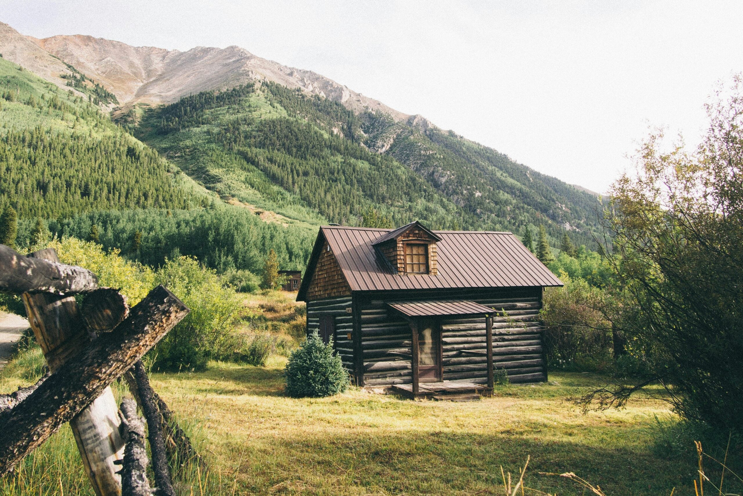 A rustic wooden cabin with a tin roof sits in a grassy clearing, surrounded by green trees and tall mountains in the background under a bright, partly cloudy sky.