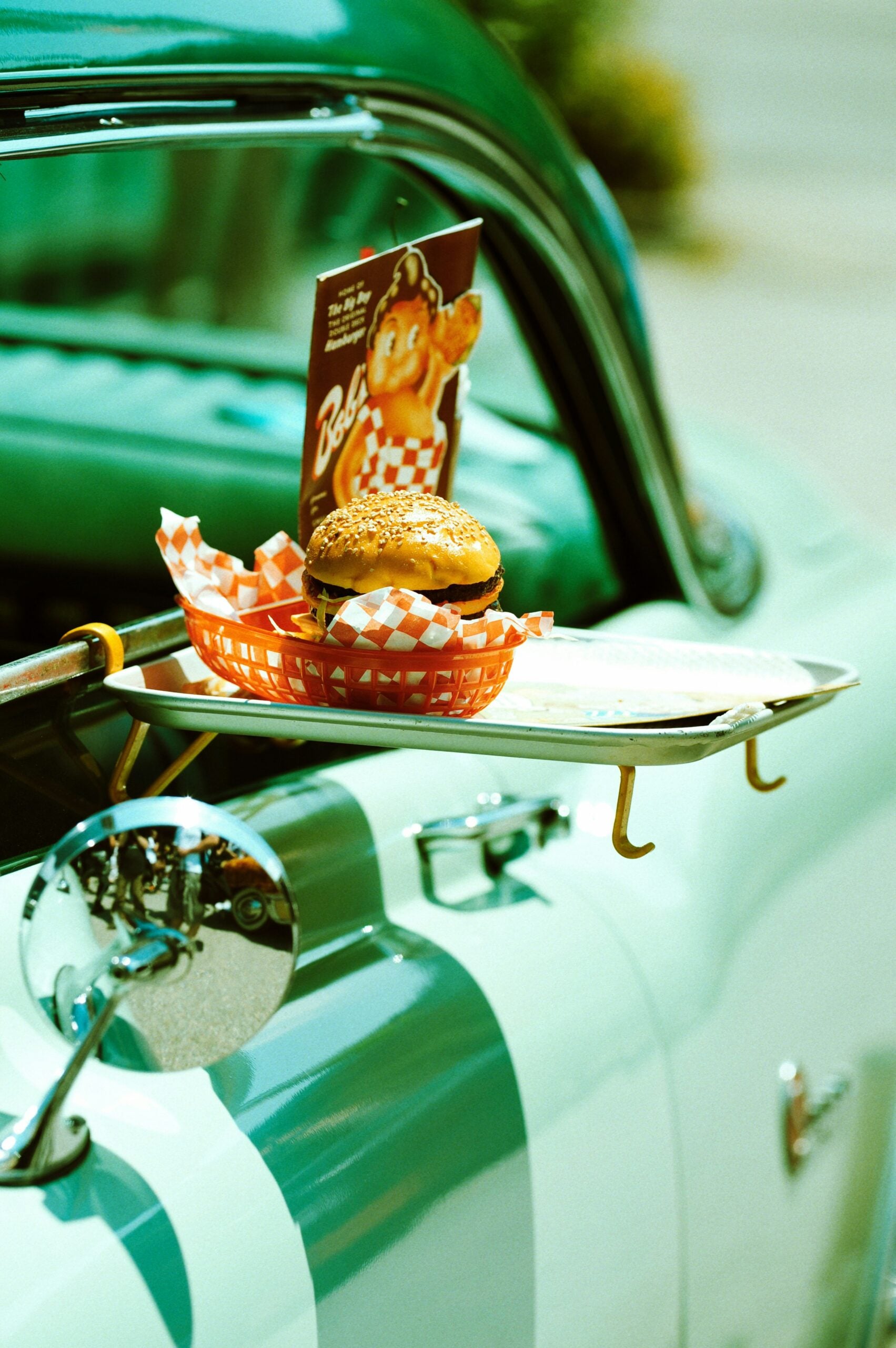 A burger in a red basket with checkered paper, and a vintage menu sits on a metal tray attached to the window of a classic green car, evoking a retro drive-in diner scene.