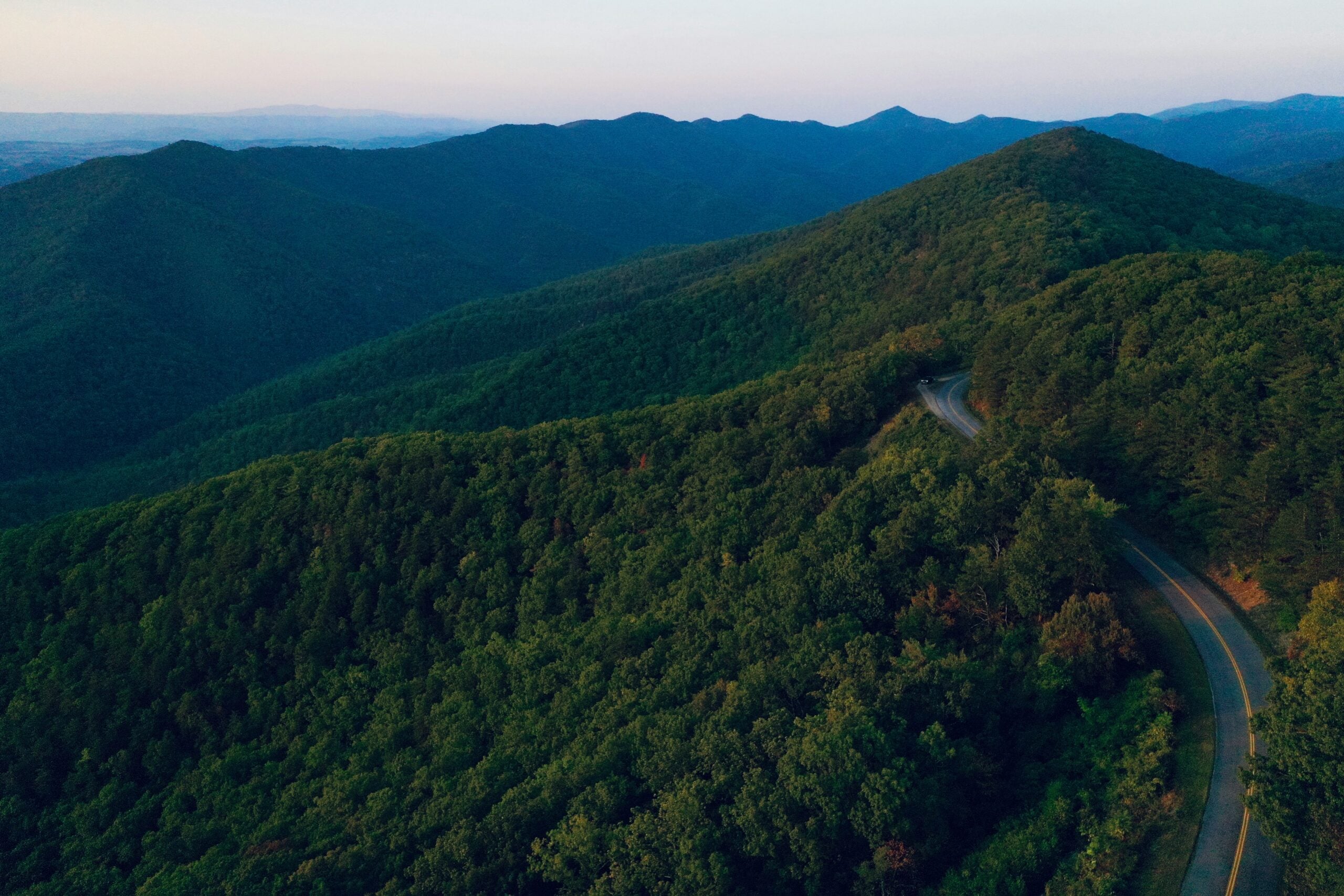 A winding road curves through lush, green, tree-covered mountains under a soft, hazy sky at sunset. The landscape stretches into the distance with layers of rolling hills.