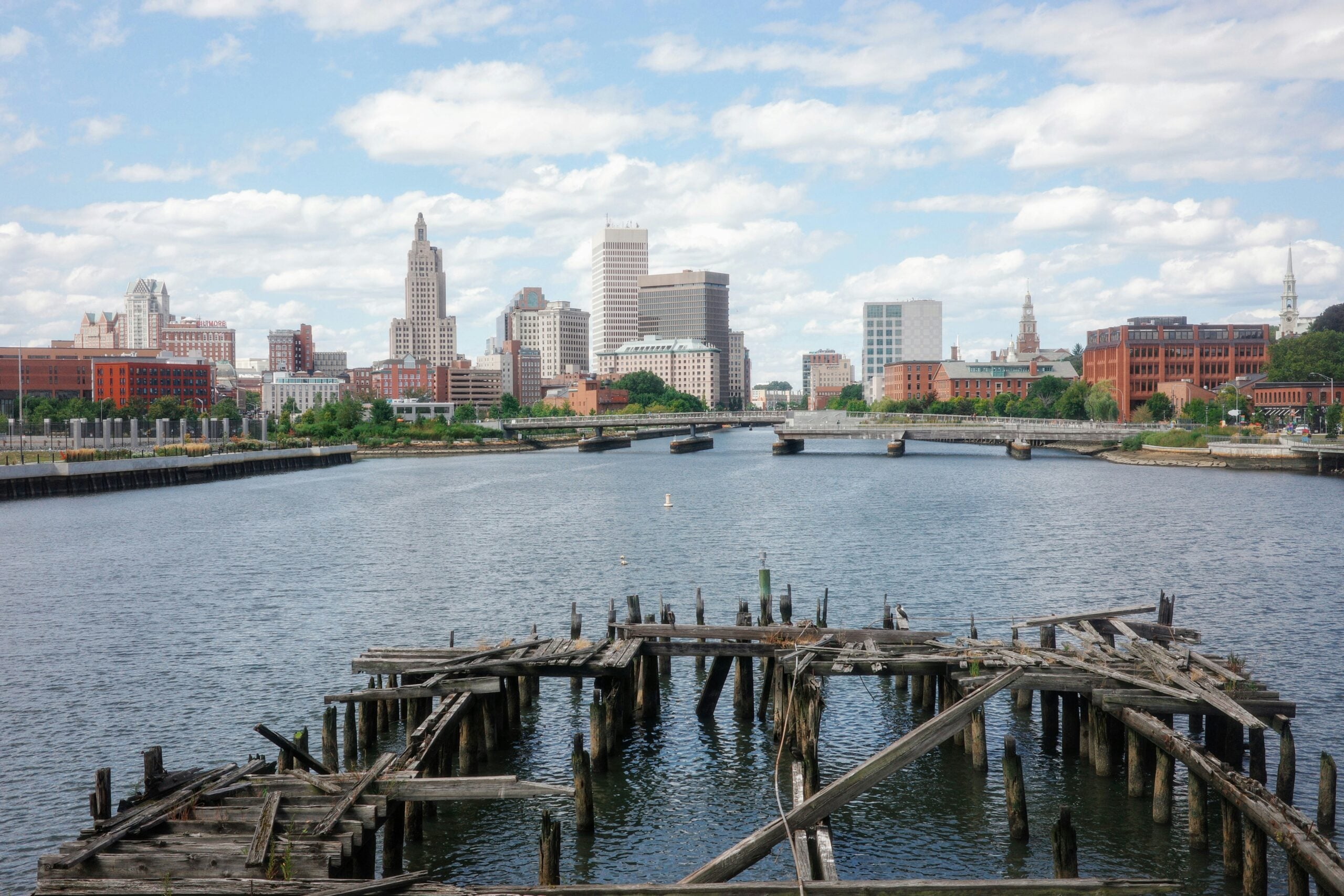 View of a river with a decaying wooden pier in the foreground and a city skyline with tall buildings and a partly cloudy sky in the background.