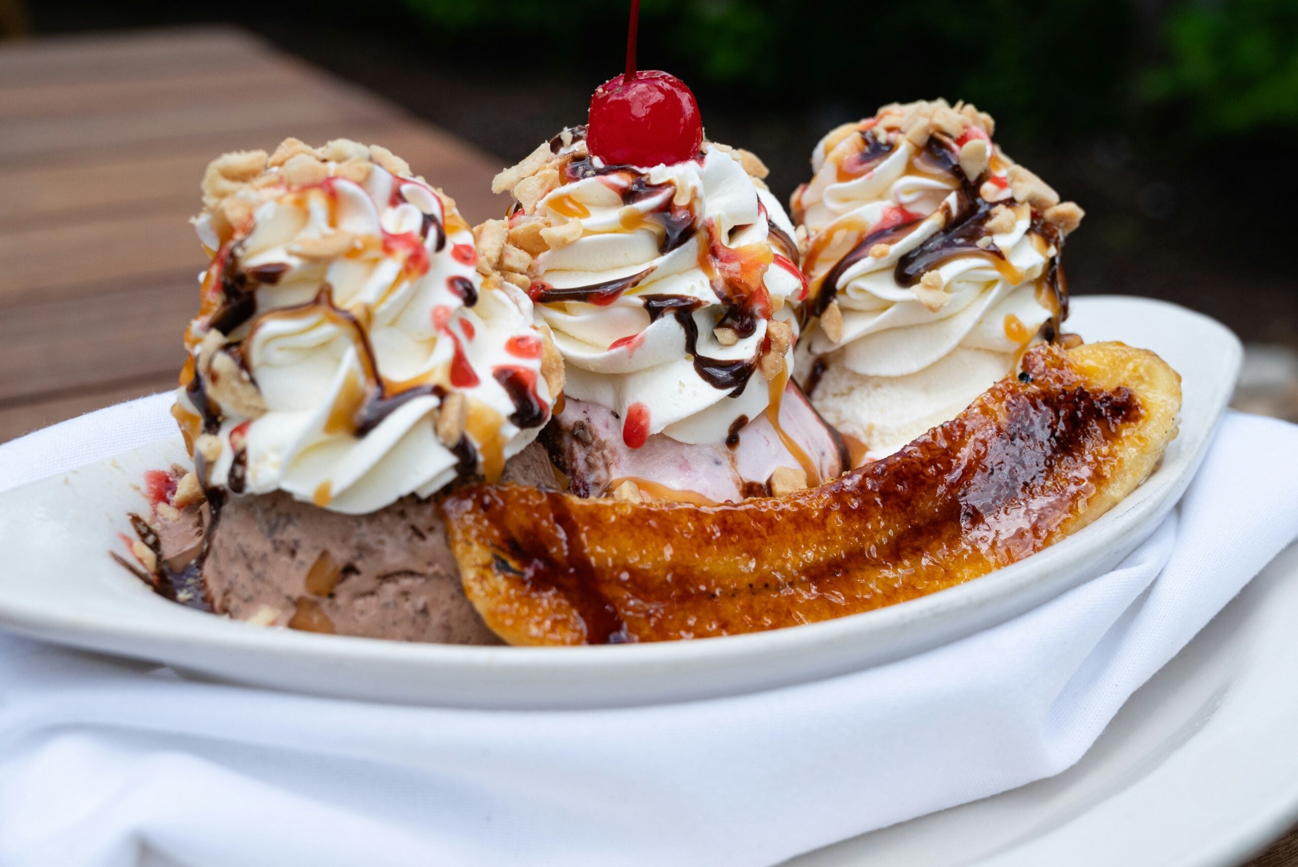 A banana split dessert with scoops of ice cream, whipped cream, chocolate and caramel drizzle, chopped nuts, a cherry on top, and a caramelized banana, served on a white plate with a white napkin underneath.