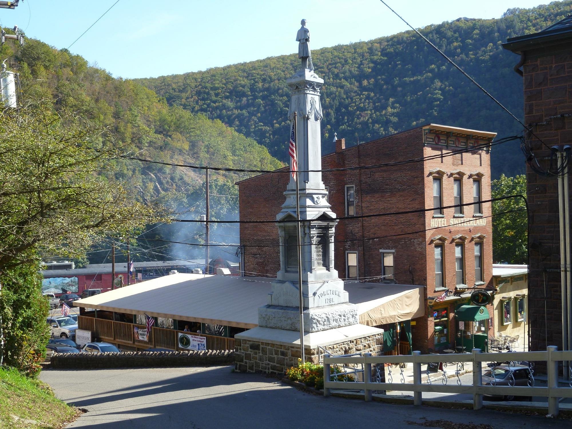 A stone soldier monument stands at a street intersection in one of the unique American towns, surrounded by historic brick buildings, trees, and distant forested hills beneath a clear sky.