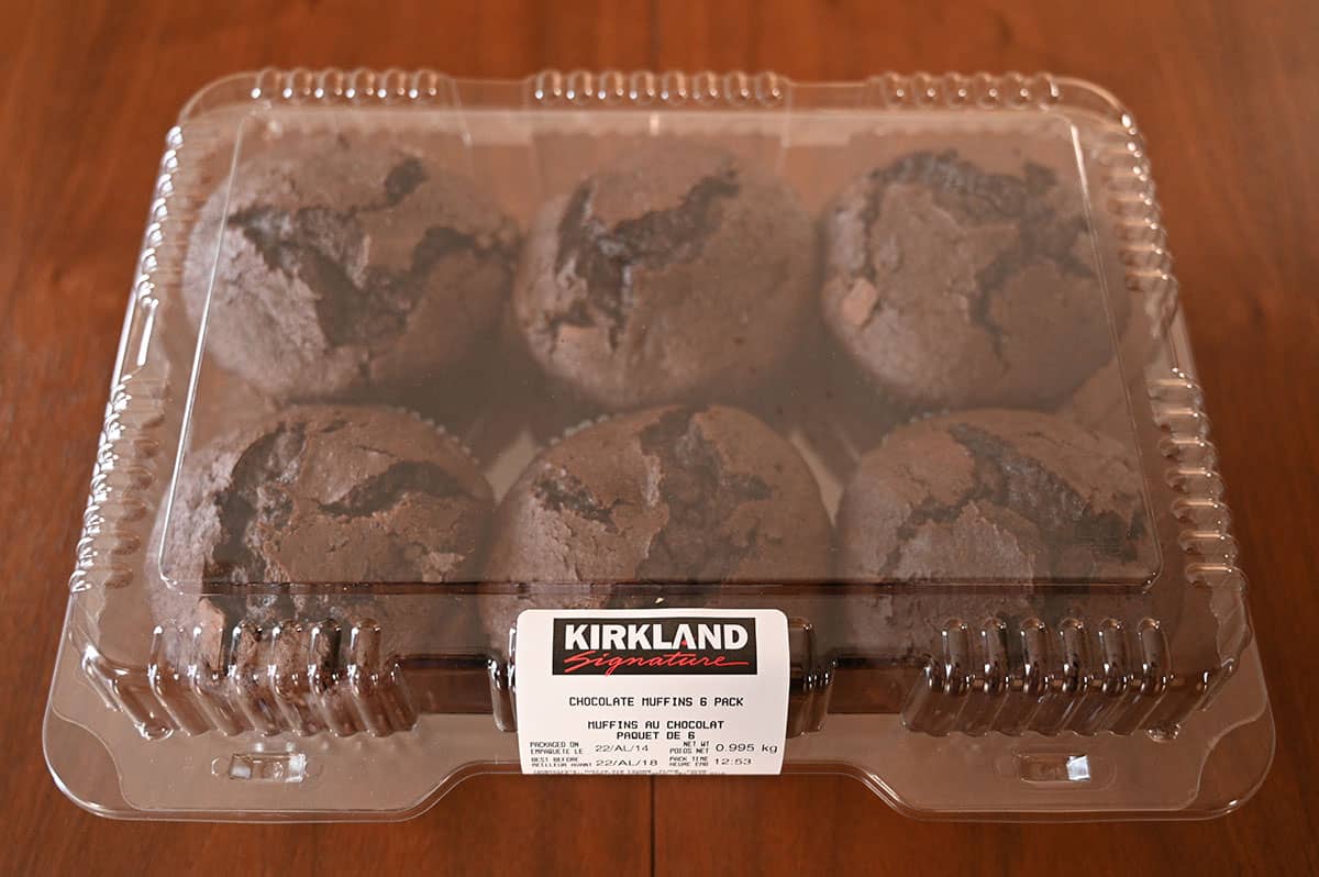 A clear plastic container holds six large chocolate muffins. The label reads "Kirkland Signature Chocolate Muffins 6 Pack" and sits on a wooden surface.