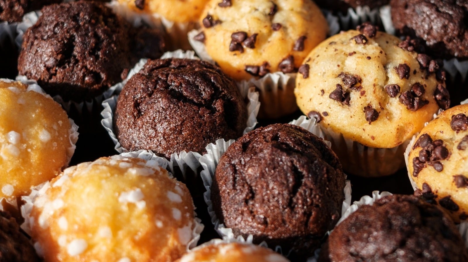 An assortment of mini muffins, including chocolate chip, chocolate, and sugar-topped varieties, arranged closely together in paper liners.