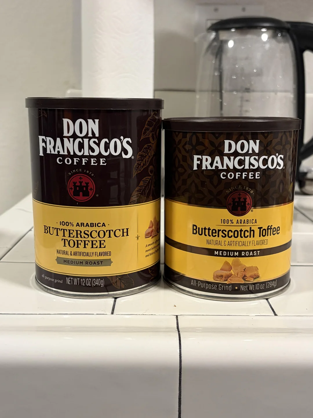 Two cans of Don Francisco’s Coffee, Butterscotch Toffee flavor, sit side by side on a white kitchen counter. The left can is 12 oz and the right can is 10 oz. A glass kettle is in the background.