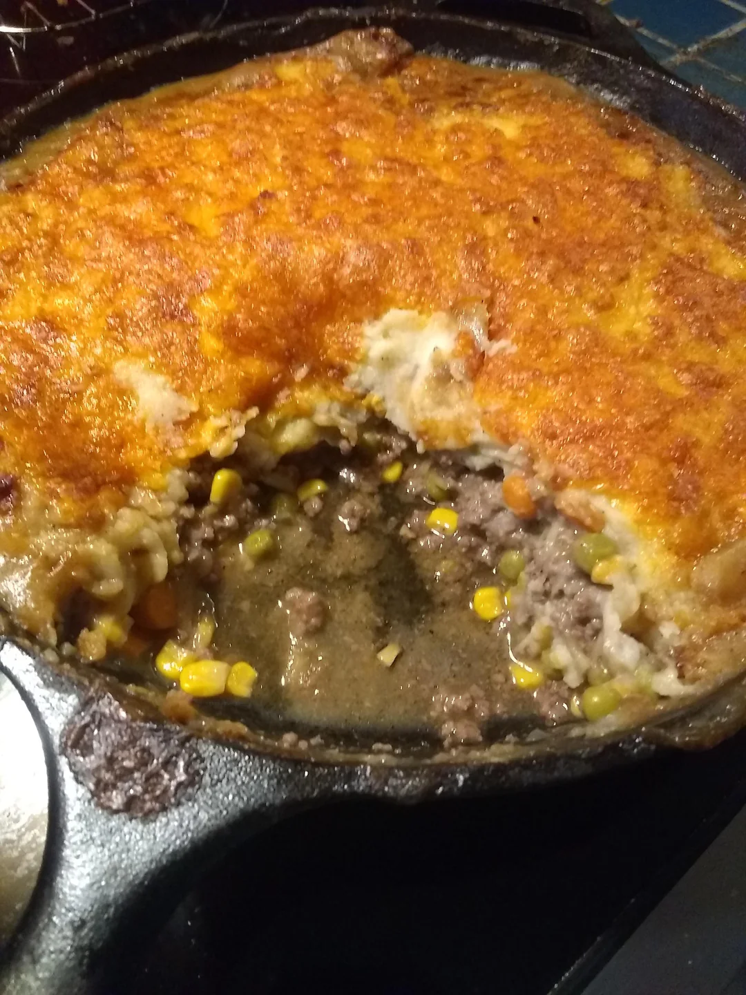 A cast iron skillet filled with shepherd’s pie, showing layers of ground beef, corn, peas, carrots, and topped with golden melted cheese and mashed potatoes. A portion has been scooped out, revealing the inside.